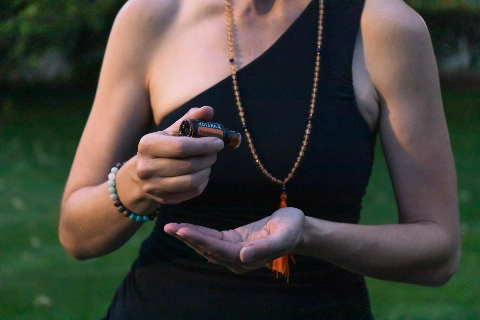 A person in a black sleeveless top holds a small amber essential oil bottle and appears to be applying or inspecting it outdoors.