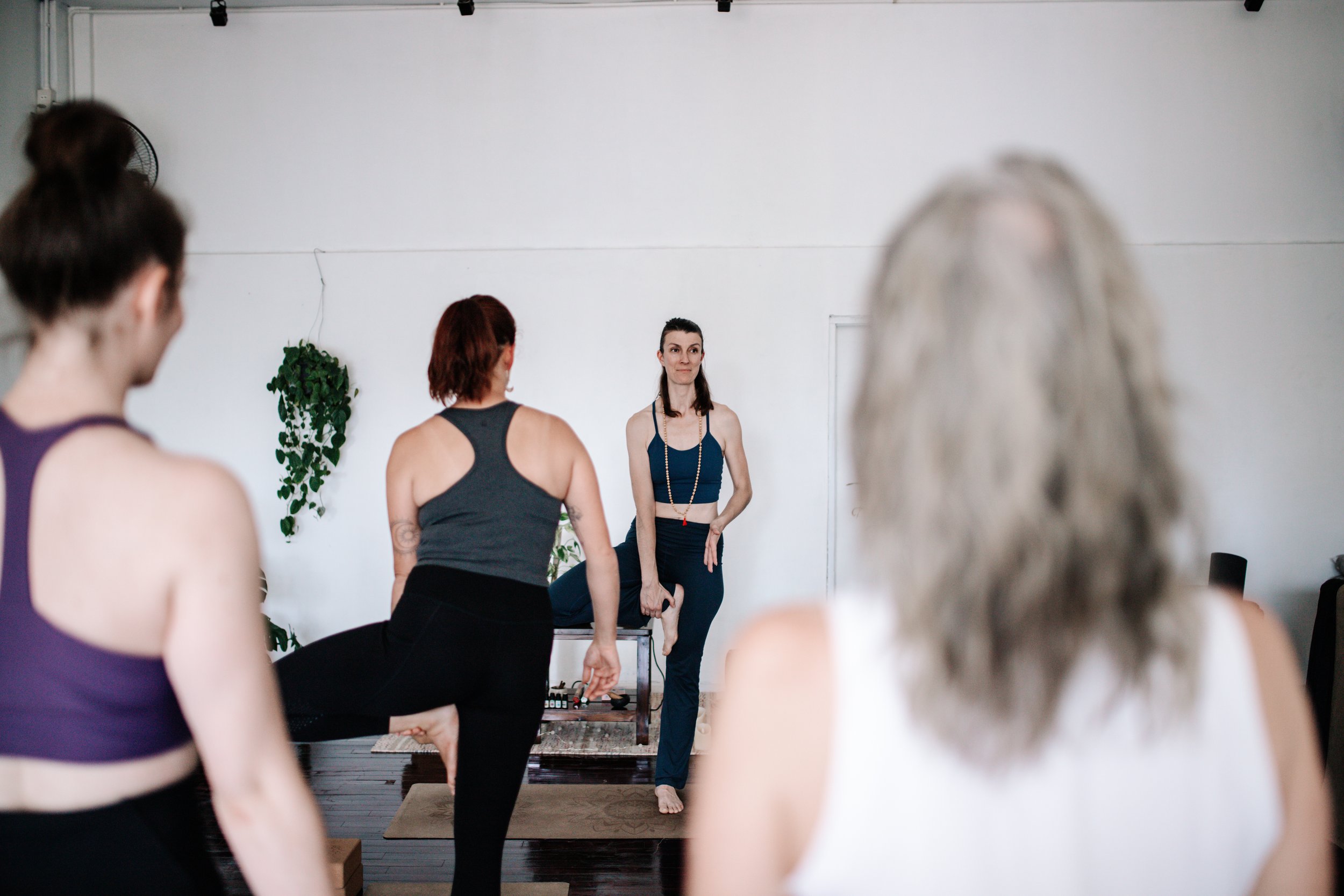 A yoga instructor leading a class in a yoga pose, with participants standing on yoga mats in a studio with white walls and plants.