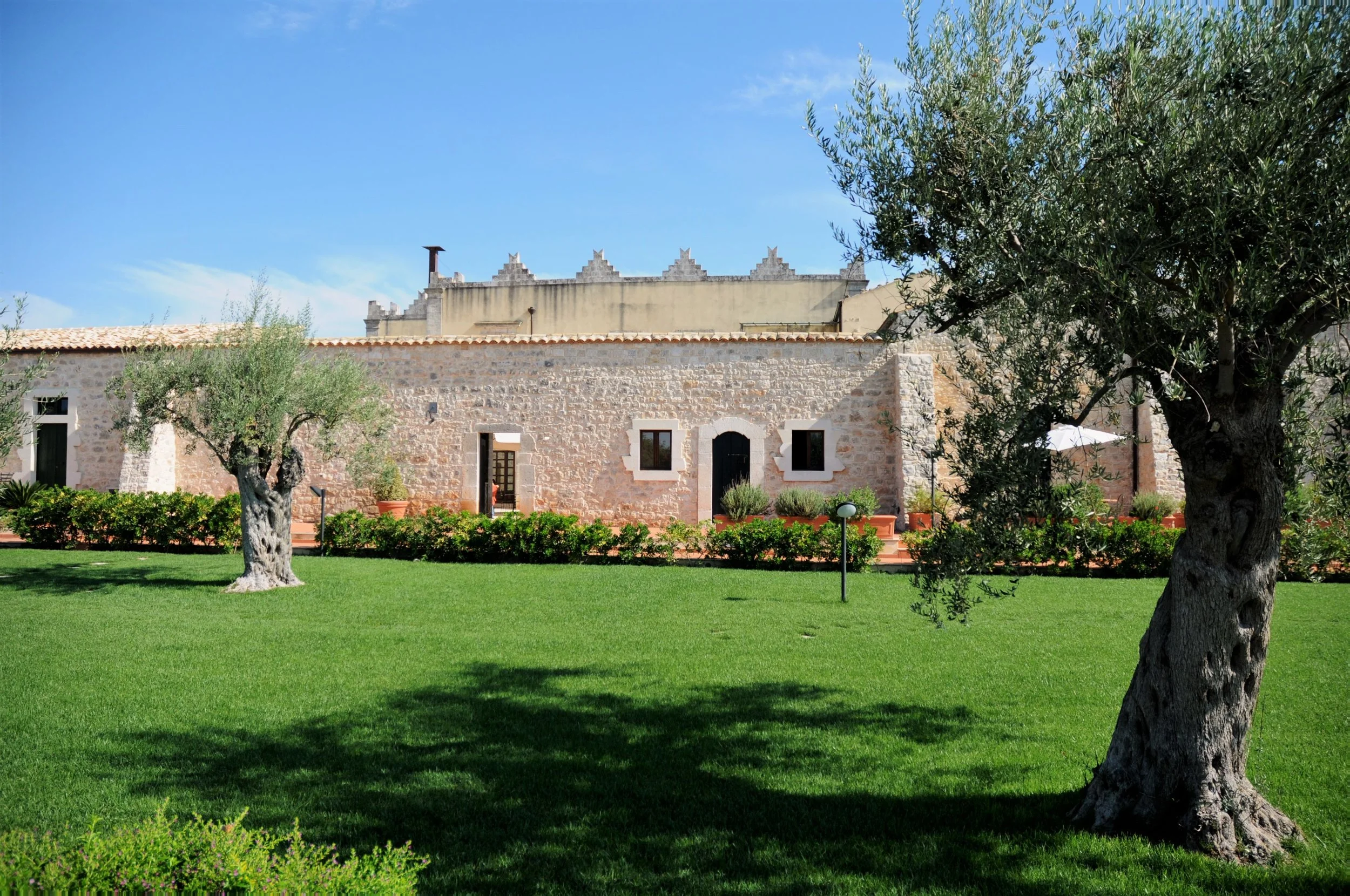 A stone house with small windows and a black door, surrounded by green grass, trees, and bushes, under a clear blue sky.