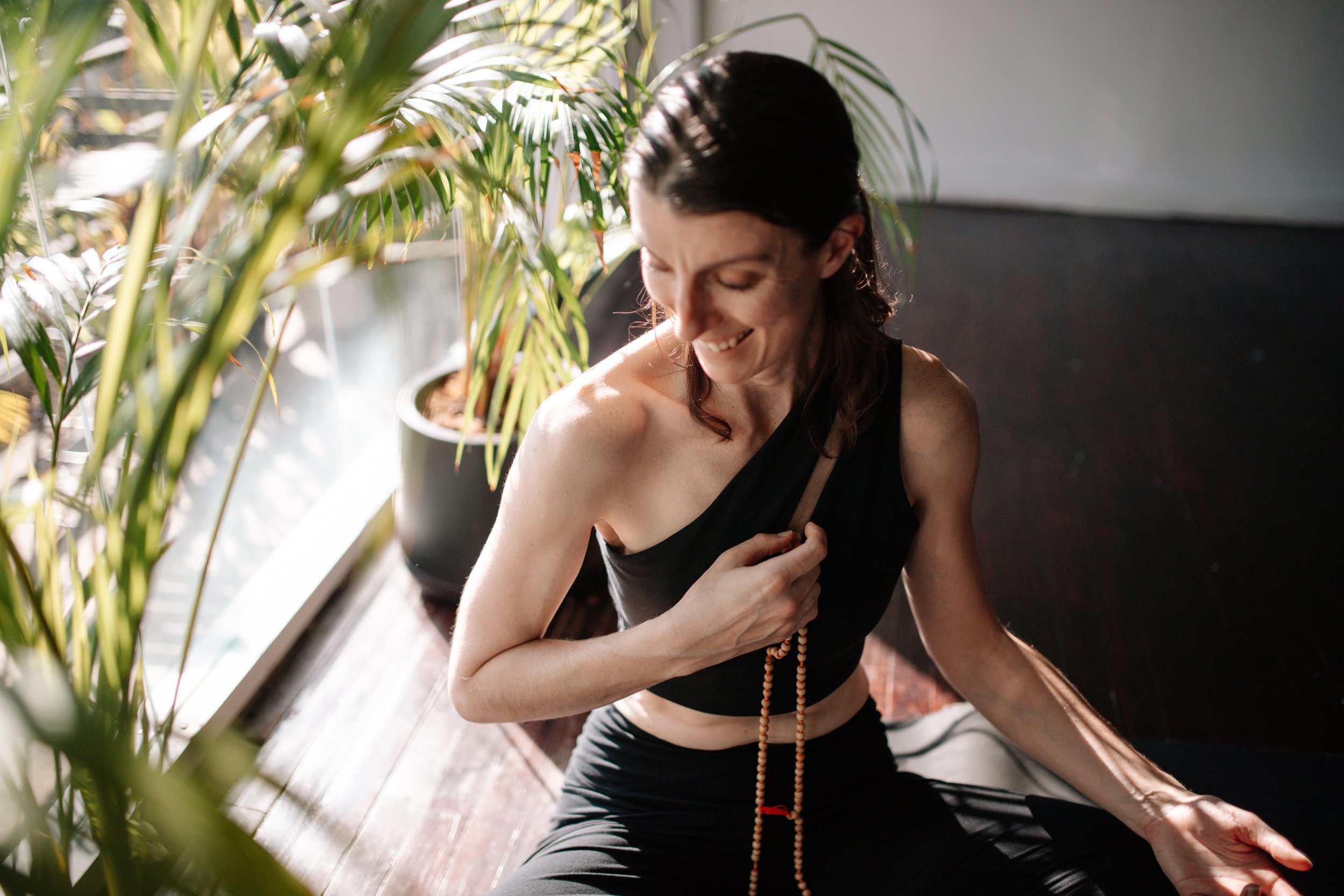 A woman with dark hair, wearing a black sleeveless top, smiling with closed eyes, sitting on the floor near a large potted plant, holding a string of beads with her left hand.