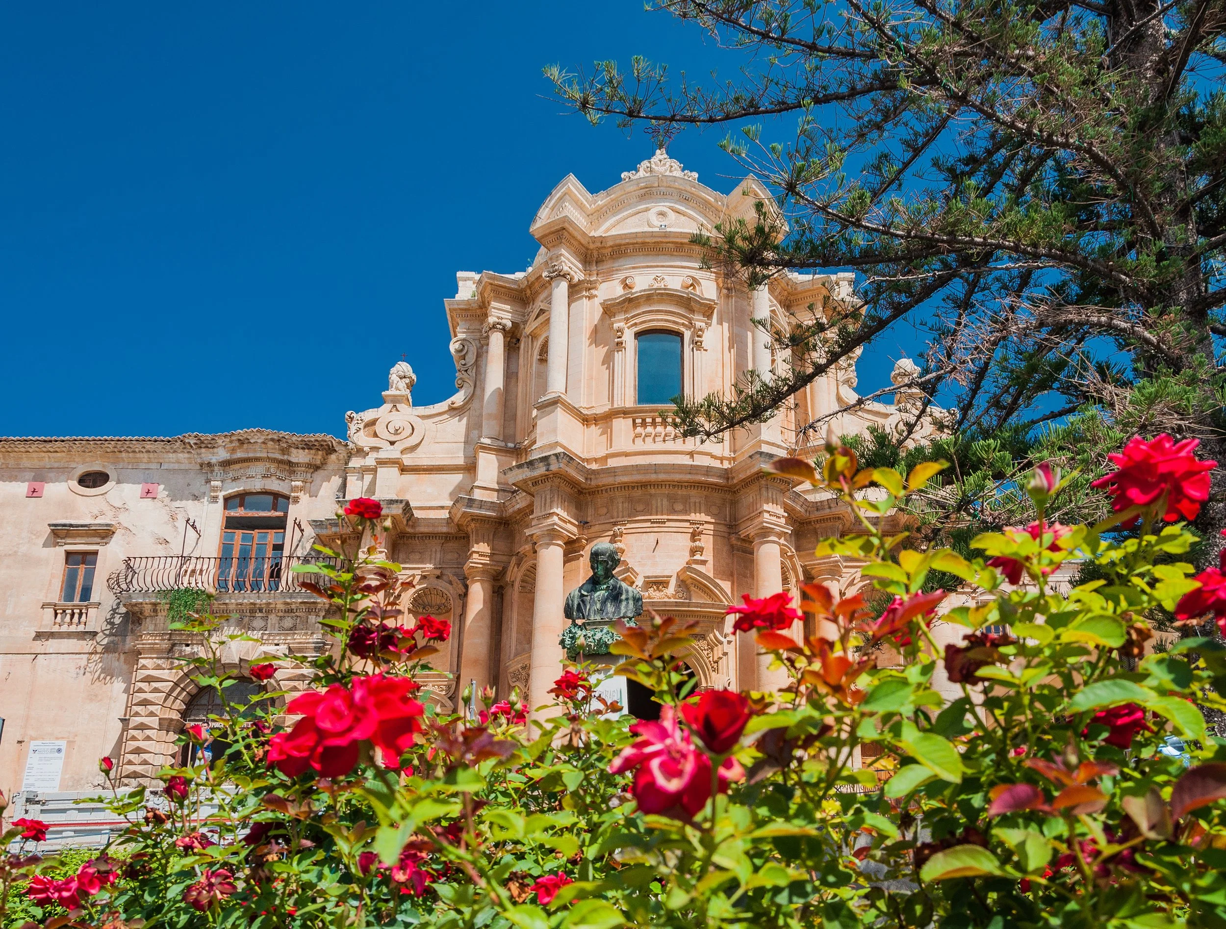 Baroque-style church with ornate facade and columns, surrounded by flowering bushes and trees, under a clear blue sky.