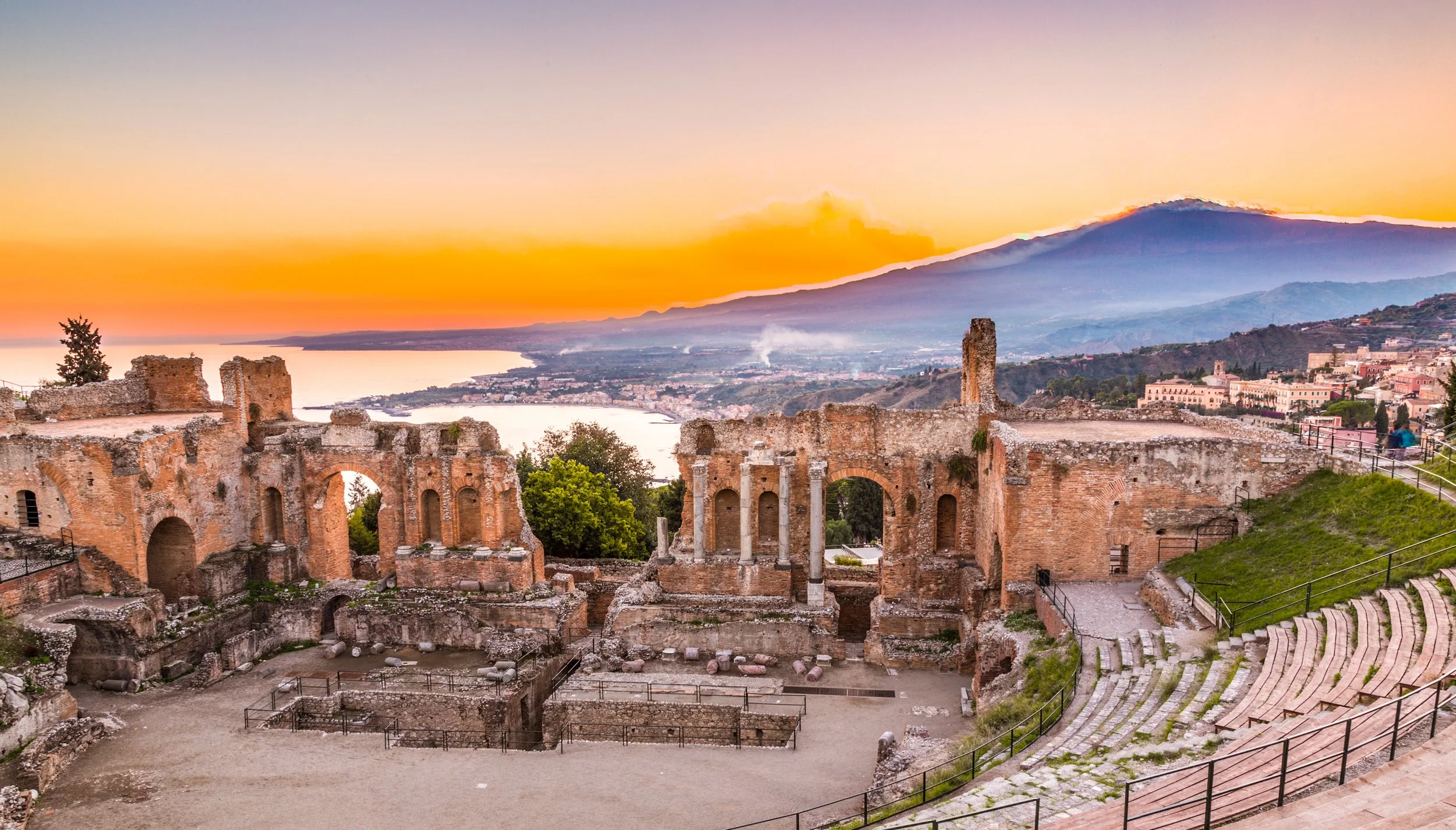 Ancient Roman ruins overlooking a body of water and a mountainous landscape during sunset.