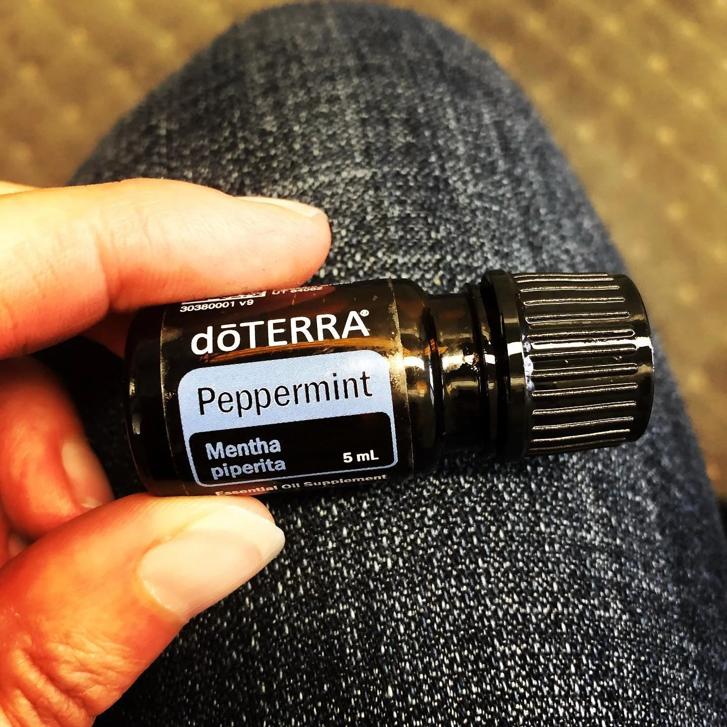 A small dark glass bottle of doTERRA Peppermint essential oil, with a black screw cap, held in a person's hand over their lap, which is covered in blue denim jeans.