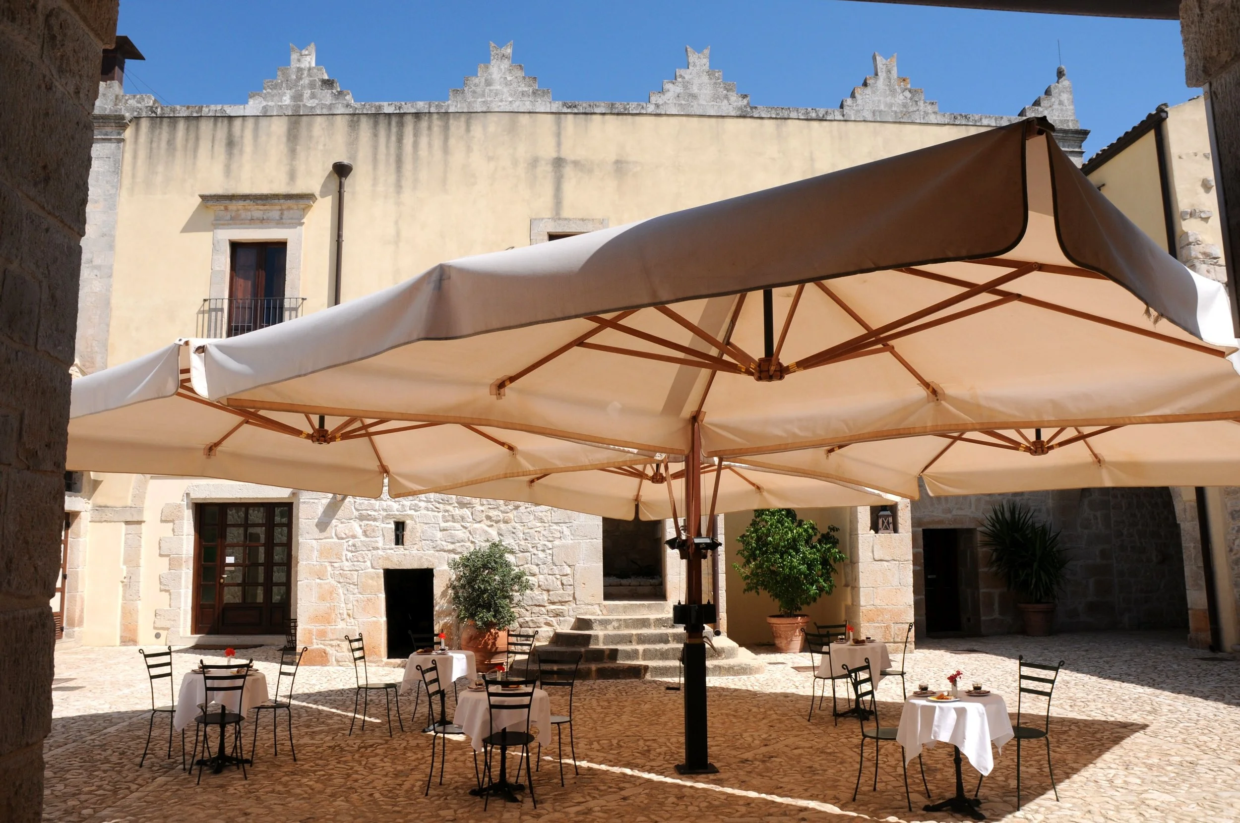 Outdoor patio with several small tables covered with white tablecloths, set with plates and cutlery, under a large beige umbrella, with stone buildings, potted plants, and a clear blue sky in the background.
