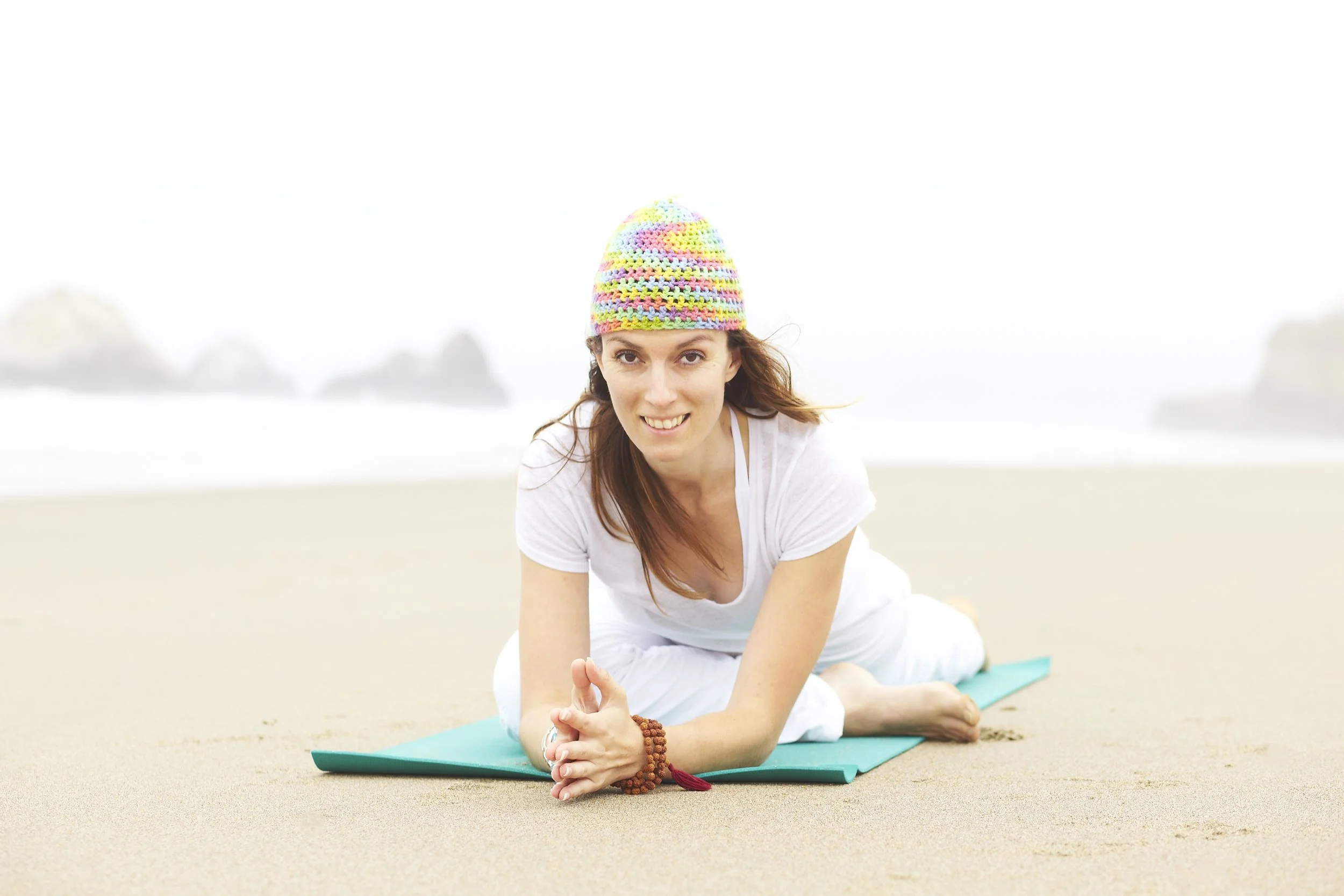 A woman in a white shirt and white pants practicing yoga on a mat on a beach, wearing a colorful knit hat and bracelets, with rocks and the ocean in the background.
