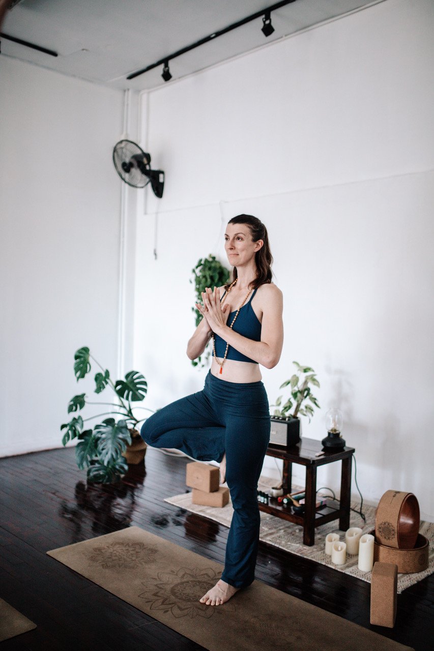 A woman practicing yoga indoors, balancing on one foot with hands in prayer position, surrounded by plants, candles, and yoga props.
