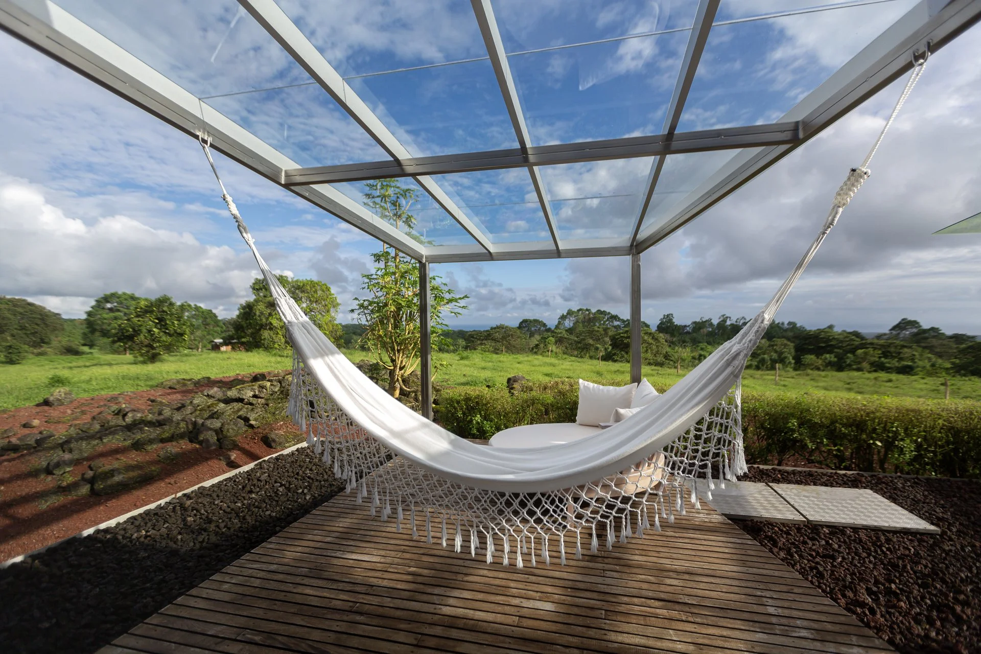 White hammock with pillows on a wooden deck under a glass roof, overlooking a green grassy landscape with trees and a partly cloudy sky.