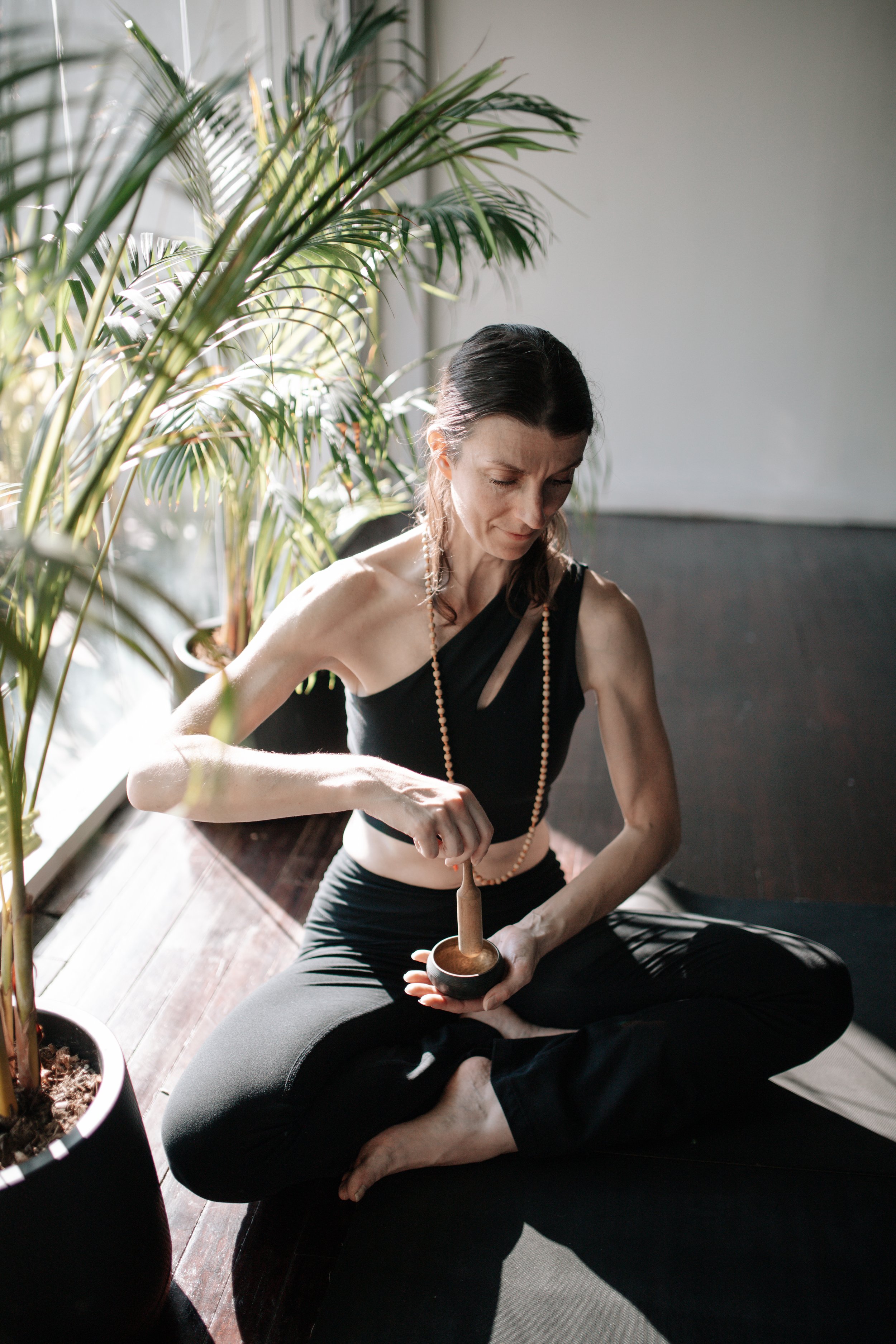Woman sitting cross-legged on a dark wooden floor, using a mortar and pestle, near large green potted plants, with sunlight streaming in through a window.