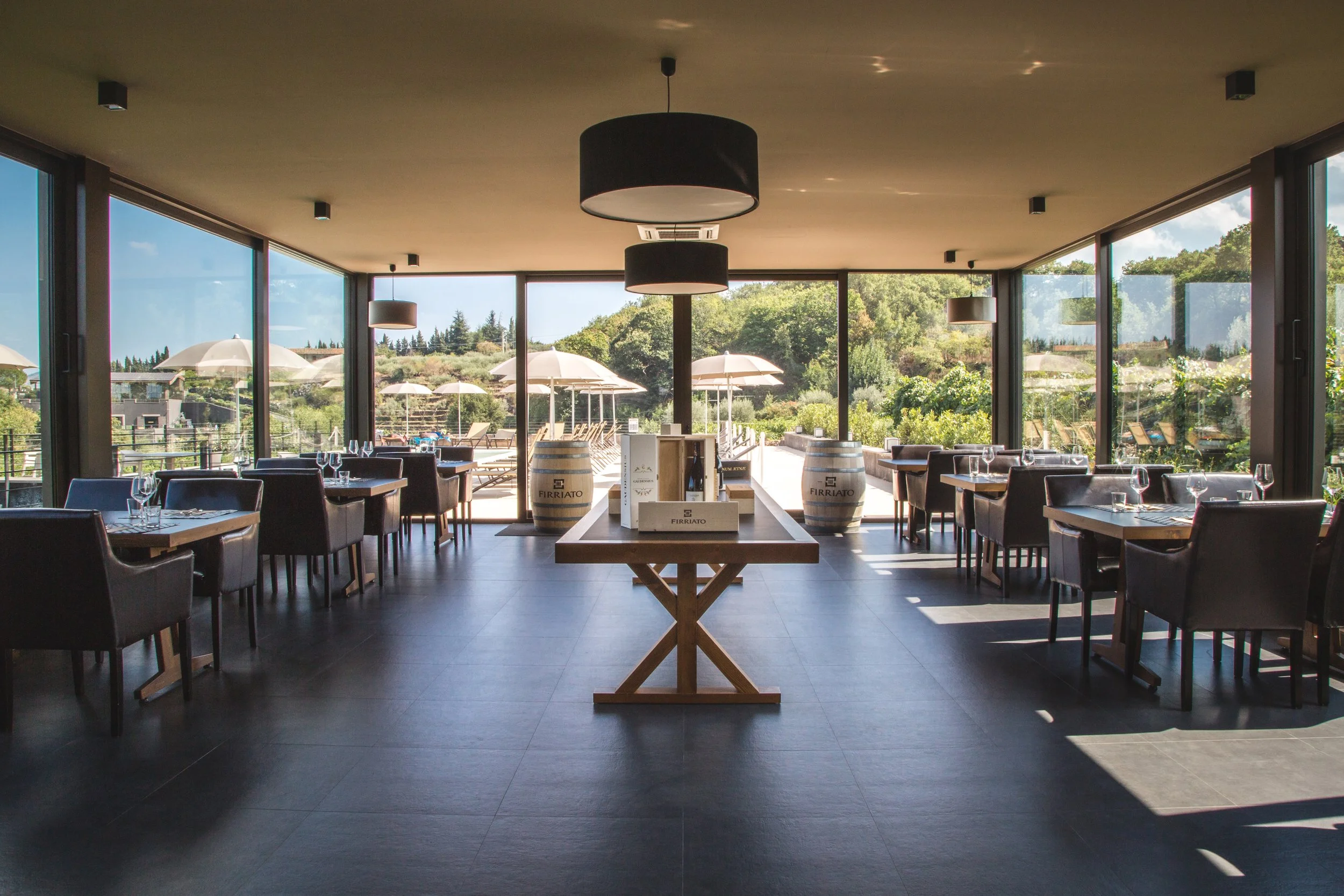 Elegant restaurant with large glass windows, outdoor views, and a central table with wine barrels and wine bottles.