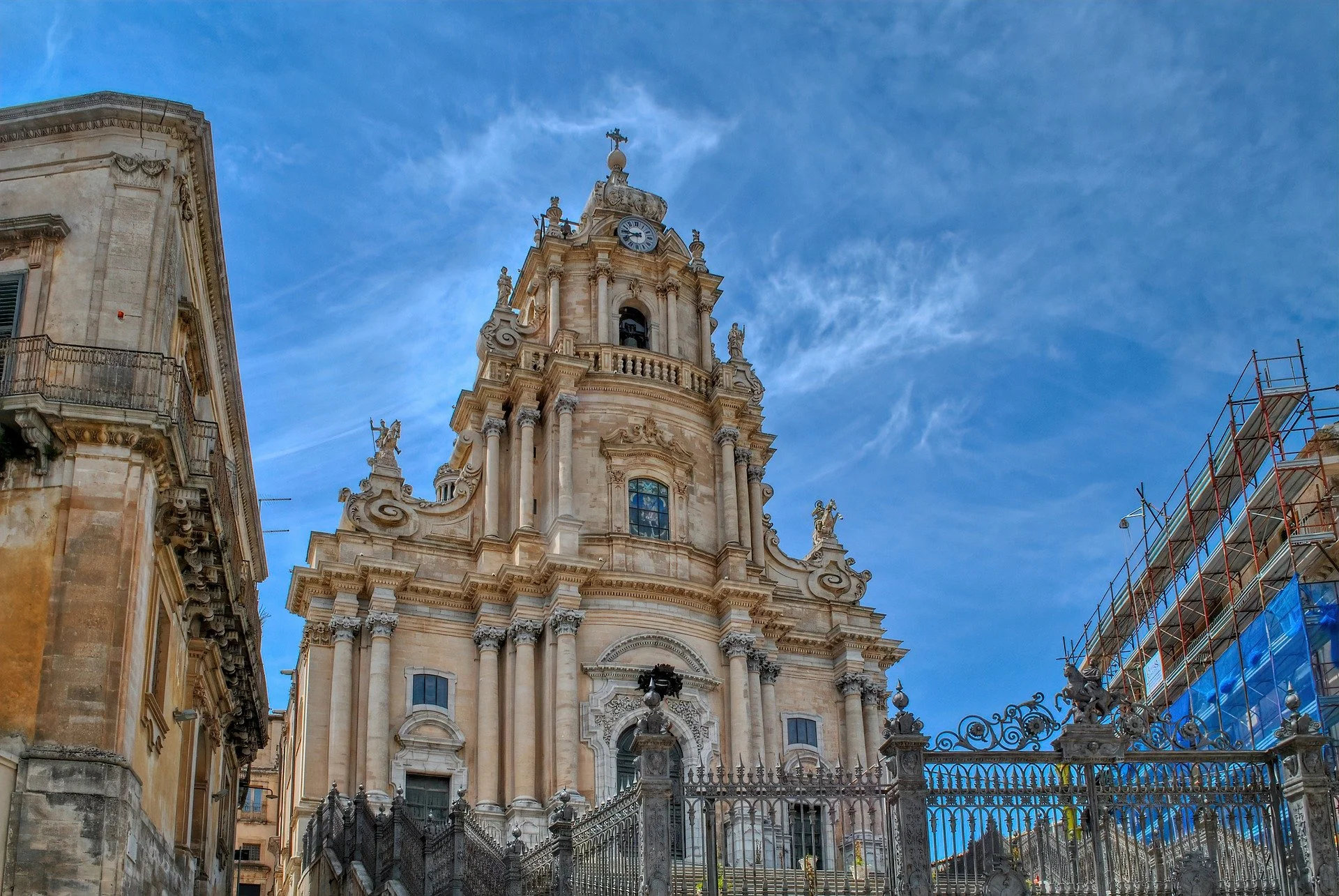 Baroque-style church with a tall ornate bell tower featuring a clock, surrounded by an iron fence, under a blue sky with wispy clouds, and scaffolding on adjacent buildings.