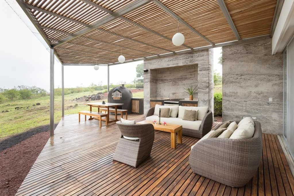 Outdoor patio with wooden flooring, wicker couches with white cushions, a wooden coffee table, a dining table with benches, a concrete wall with a potted plant, and a pizza oven in a rural setting.