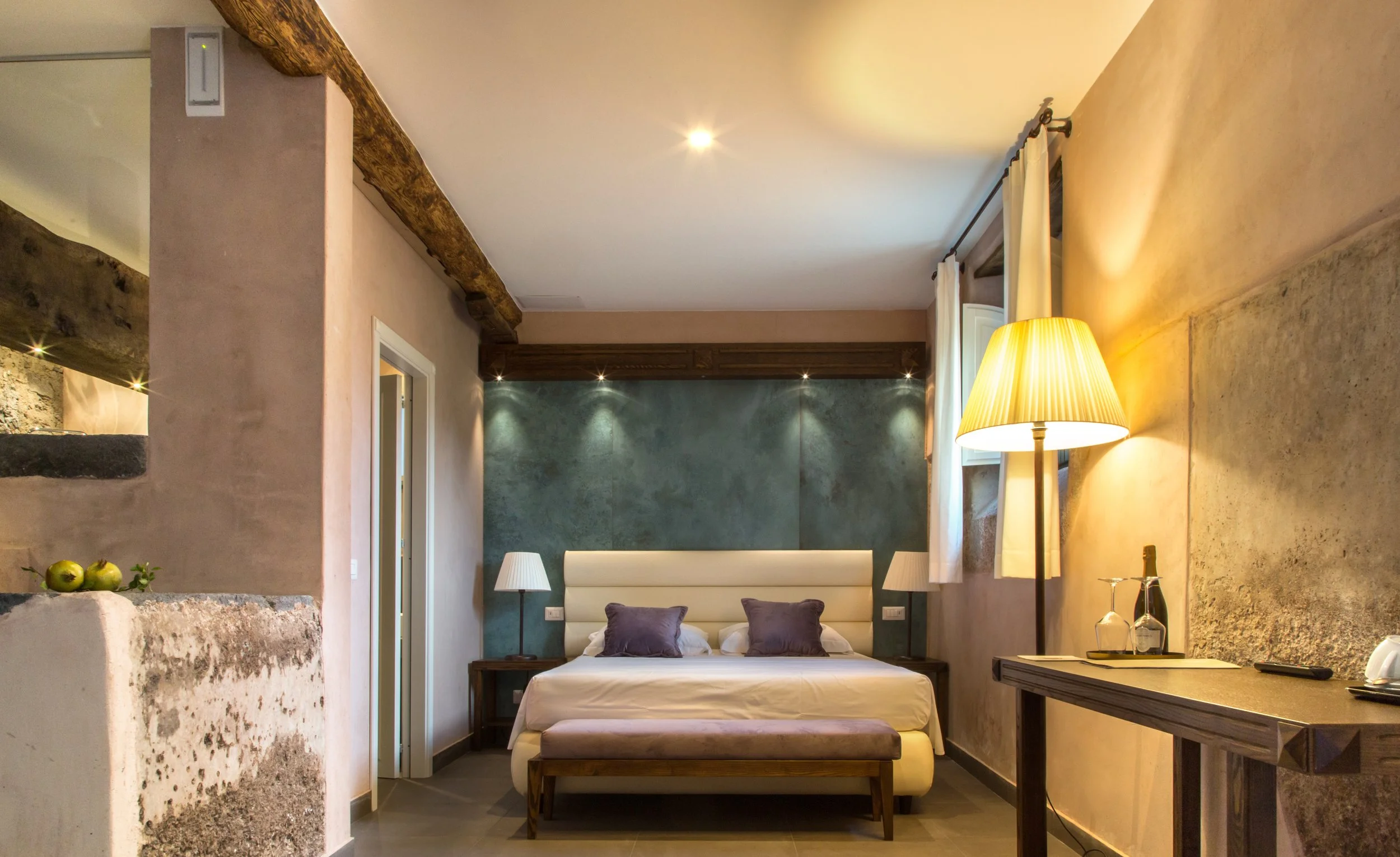 A cozy bedroom with a large bed featuring a white headboard, two bedside lamps, a floor lamp, and a window with white curtains. The room has textured walls and wooden beams on the ceiling.