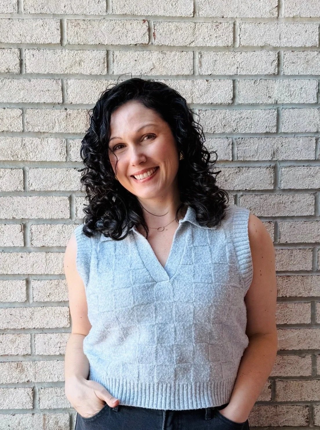 A woman with curly black hair wearing a light blue summer-sweater, standing in front of a white brick wall, smiling and posing with both hands resting casually in her jean pockets.