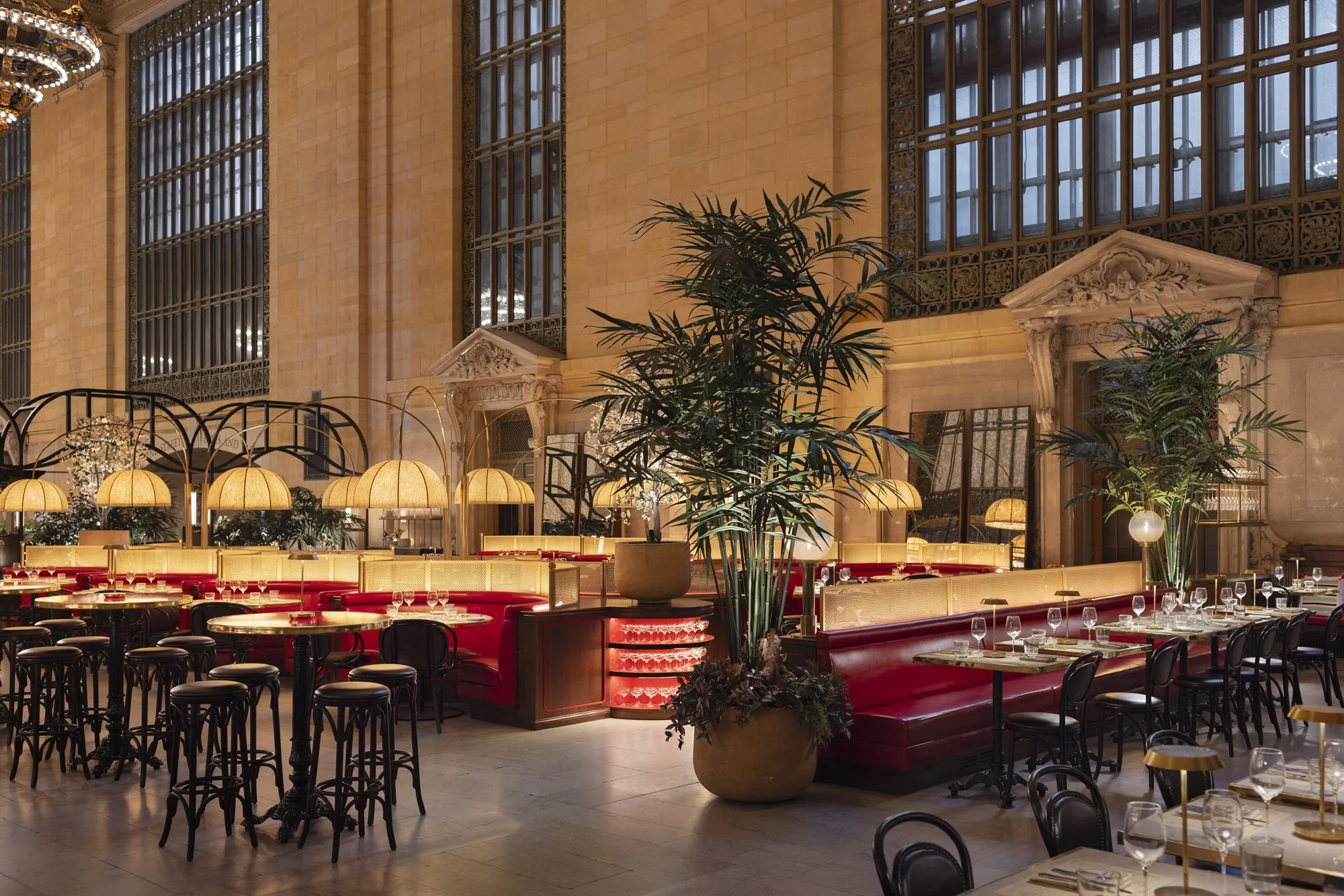 Elegant restaurant interior with large stained glass windows, tall green plants, and warmly lit tables and booths.