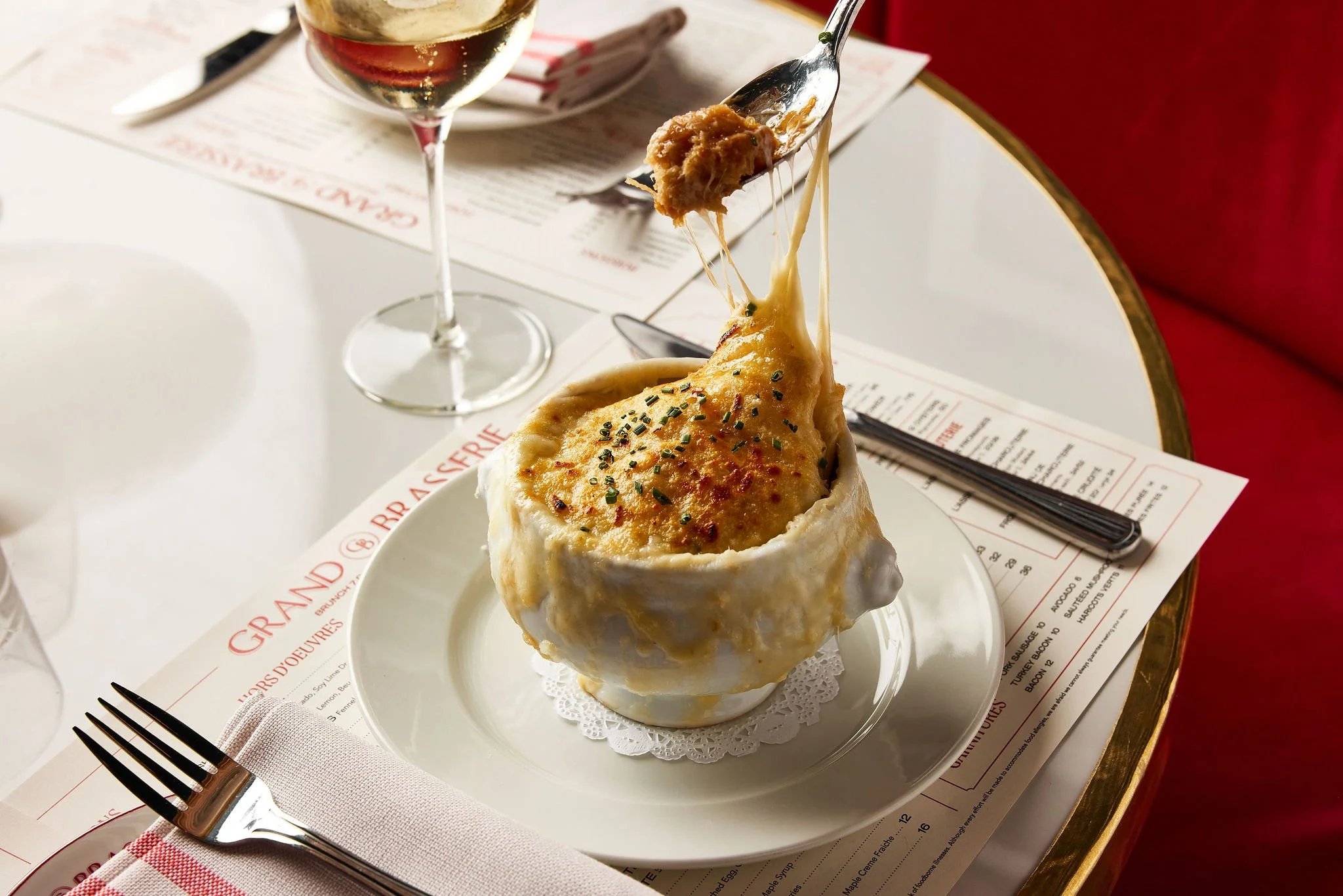 French onion soup in a bread bowl with a slice of bread topped with melted cheese, served on a white plate, with a glass of white wine and a fork on a place setting at a restaurant.