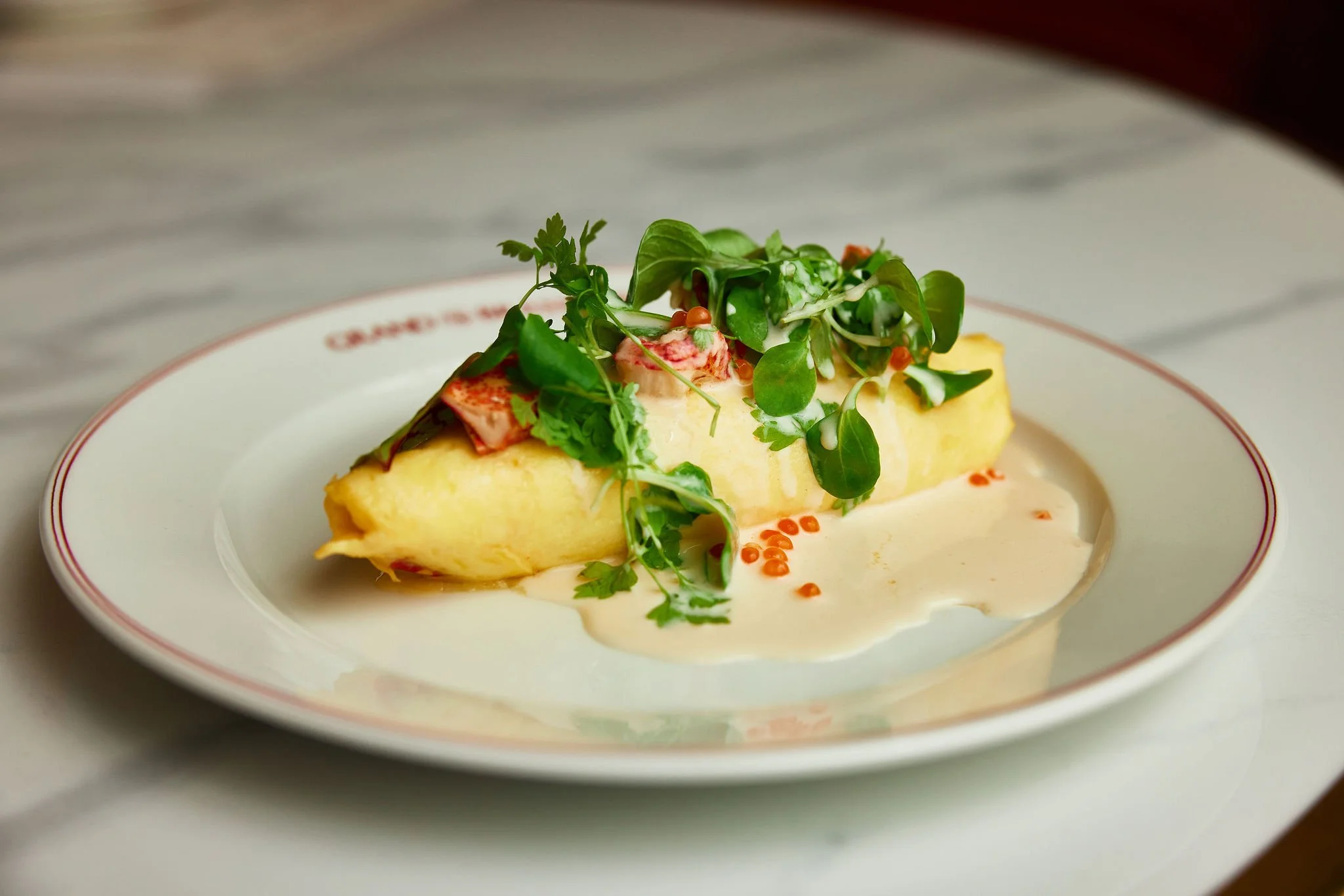 A plated dish with a rolled omelette topped with microgreens, red caviar, and sauce garnishes on a white plate with red trim.