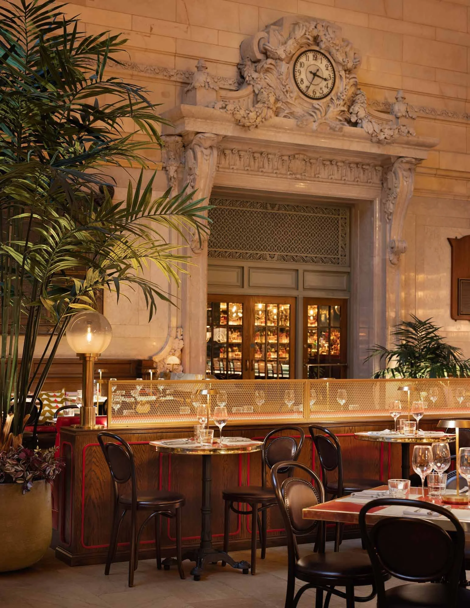 Elegant restaurant interior with a marble fireplace, ornate clock, large potted plants, and tables set with glassware and cutlery.