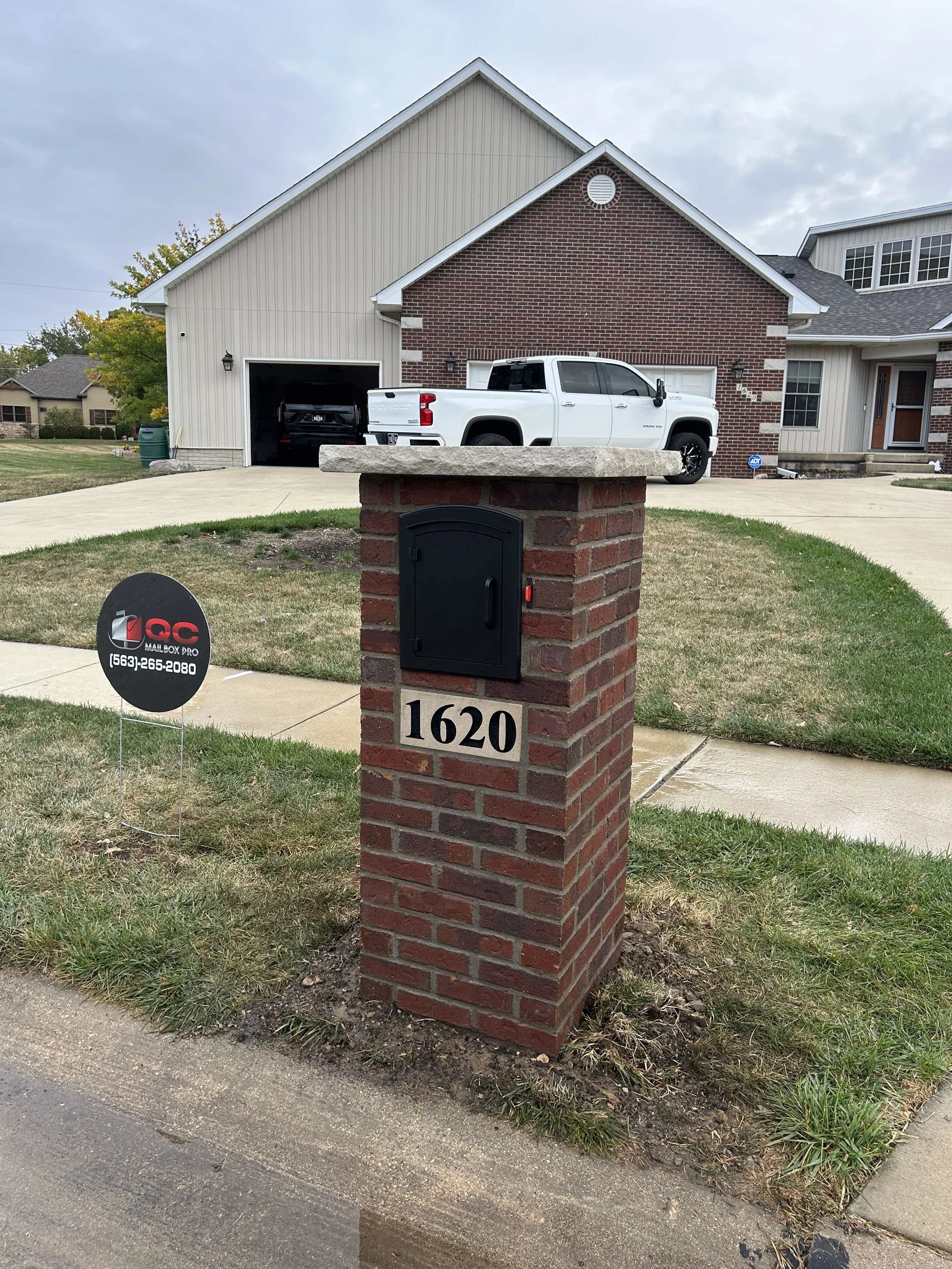 A brick mailbox with the house number 1620 in front of a suburban house. The house has a garage with a white truck parked on the driveway. There is a small sign for a mailbox business on the grass near the curb.