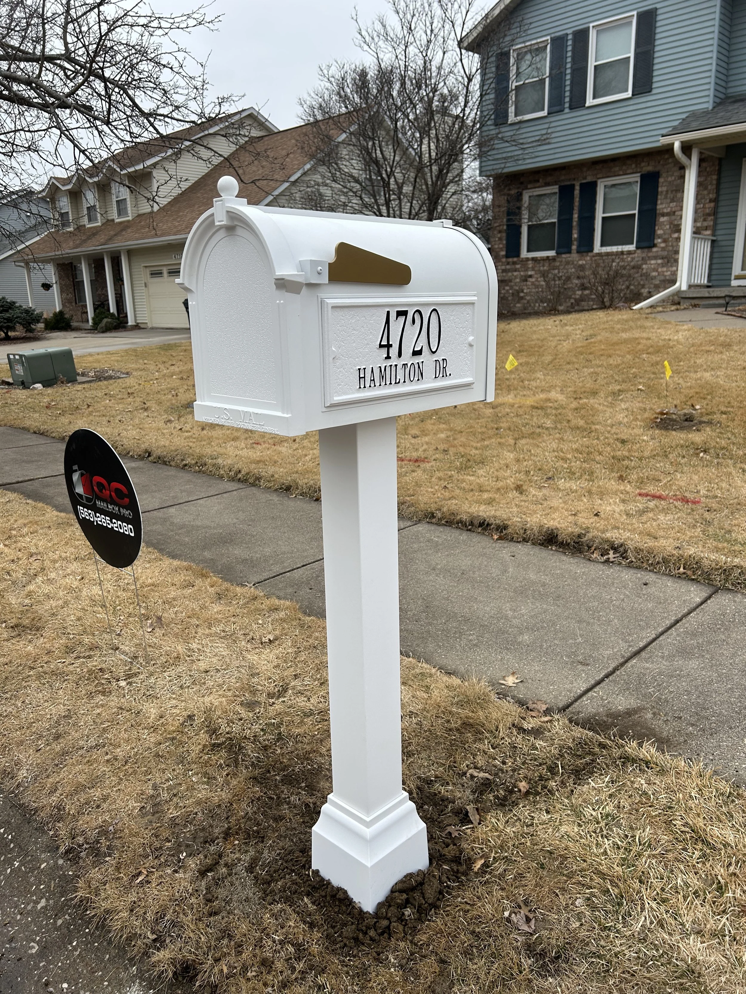 A white mailbox on a residential lawn with the address 4720 Hamilton Dr. The mailbox has a gold-colored flag and is mounted on a white post. There are neighboring houses and a sidewalk nearby.