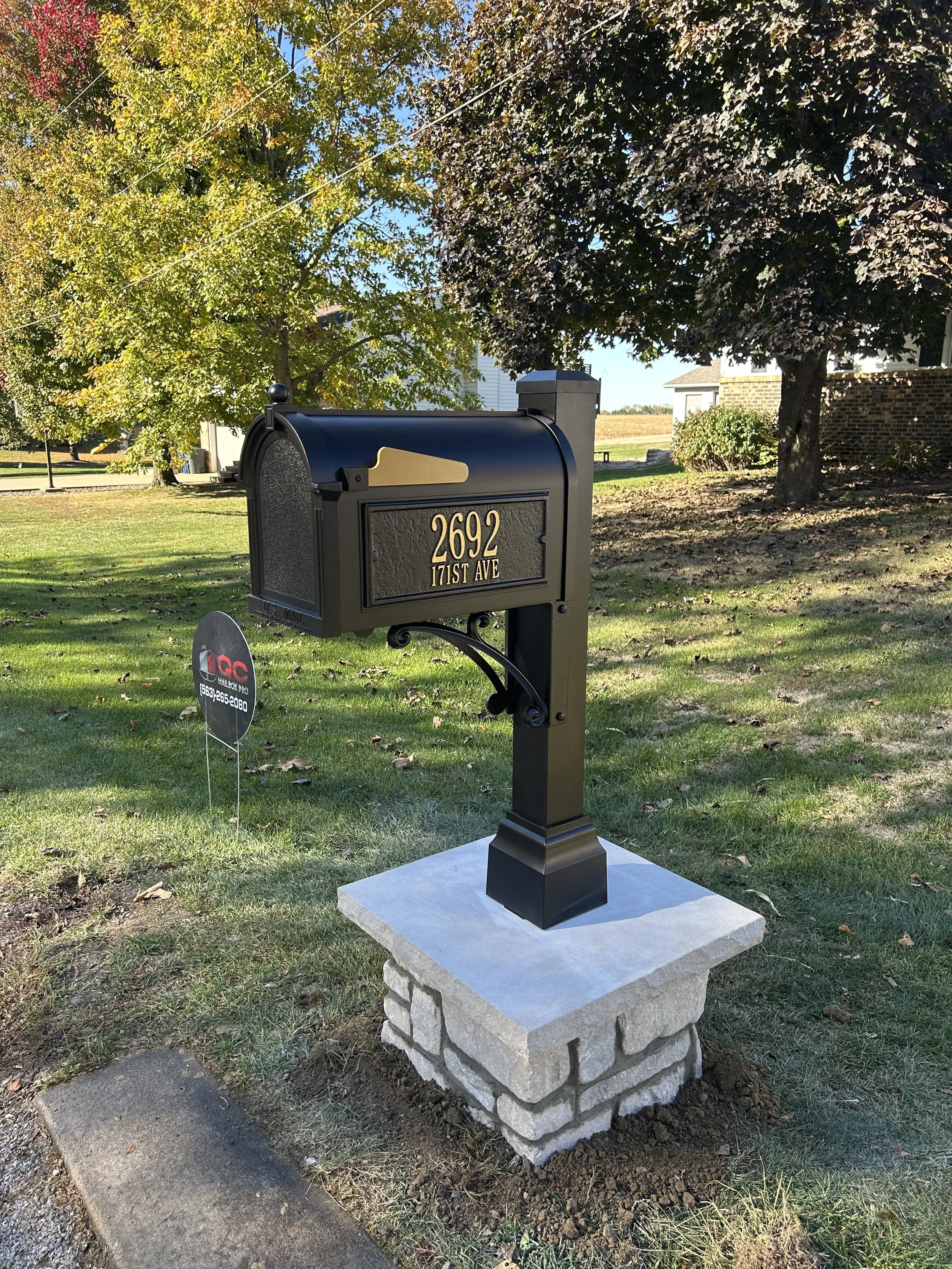 Black mailbox with gold numbers '2692' and street address '171st Ave' on a black post, mounted on a stone base in a yard with green grass and trees.