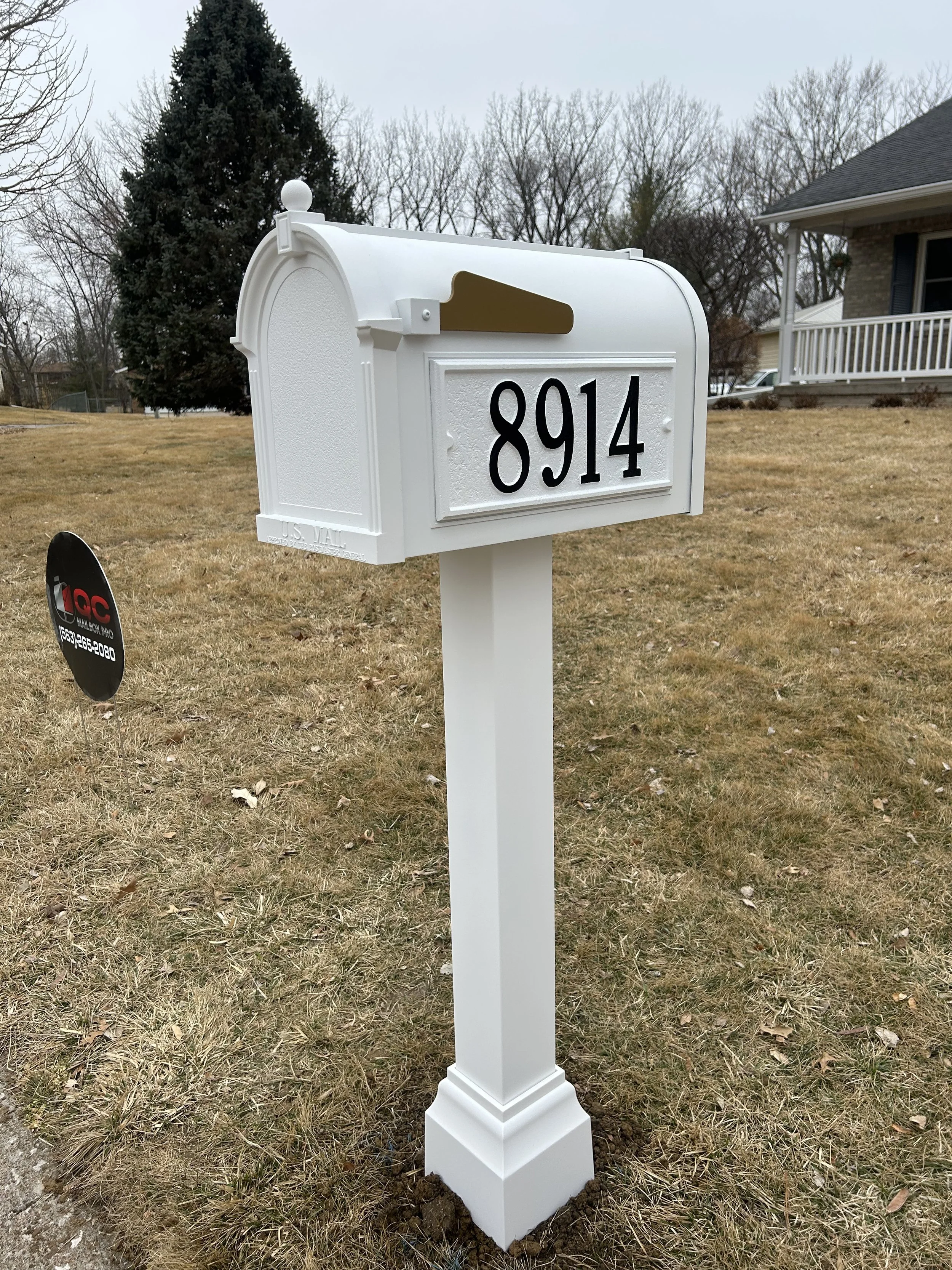 White mailbox with the house number 8914 on a white post in a front yard with grass, a large pine tree, and a house with a porch in the background.