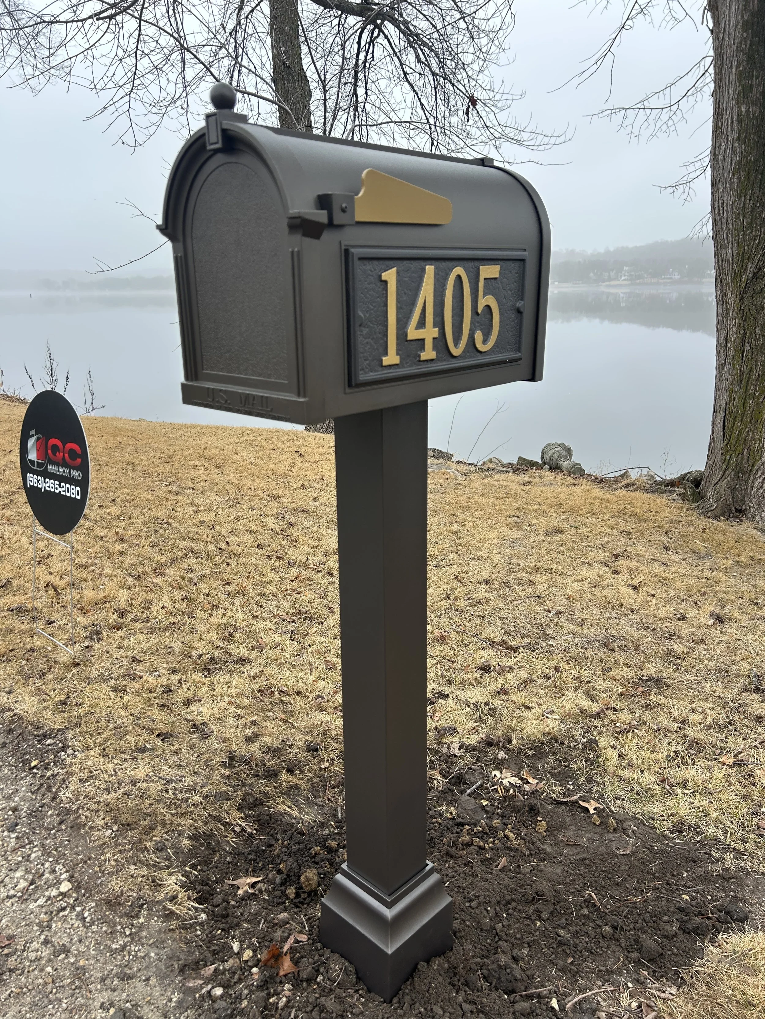 Black mailbox with gold numbers 1405 on a black post near a lake with leafless trees and overcast sky.