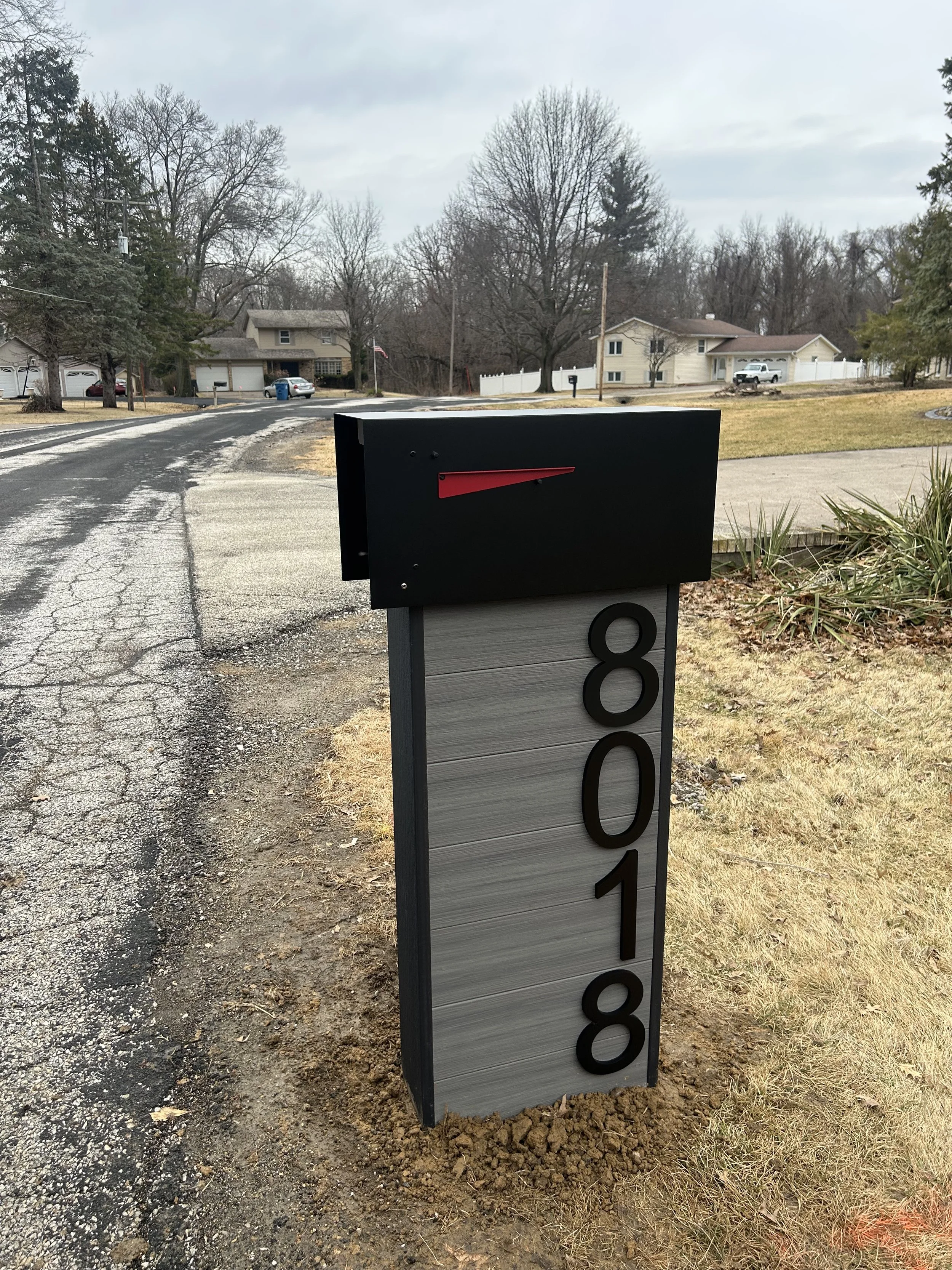 A black mailbox with a red flag and a modern house number post displaying the address 8018, situated on a lawn next to a residential street in a suburban neighborhood.
