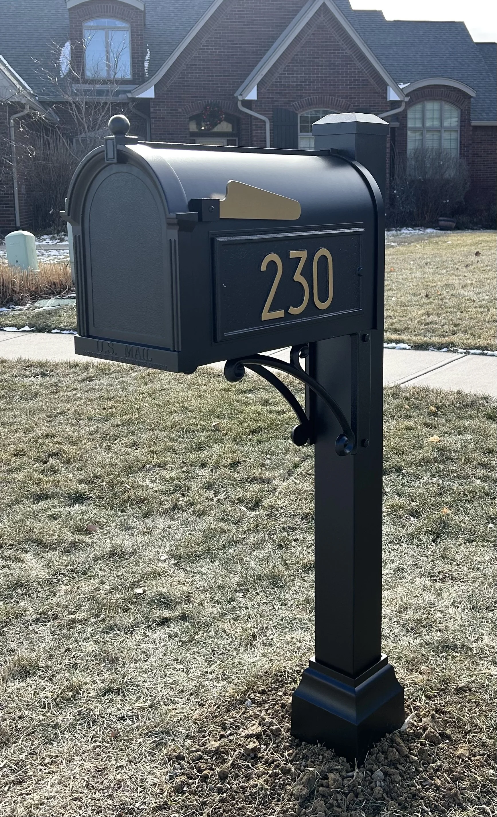 Black curbside mailbox with gold details on a lawn in front of a brick house, marked with the number 230.