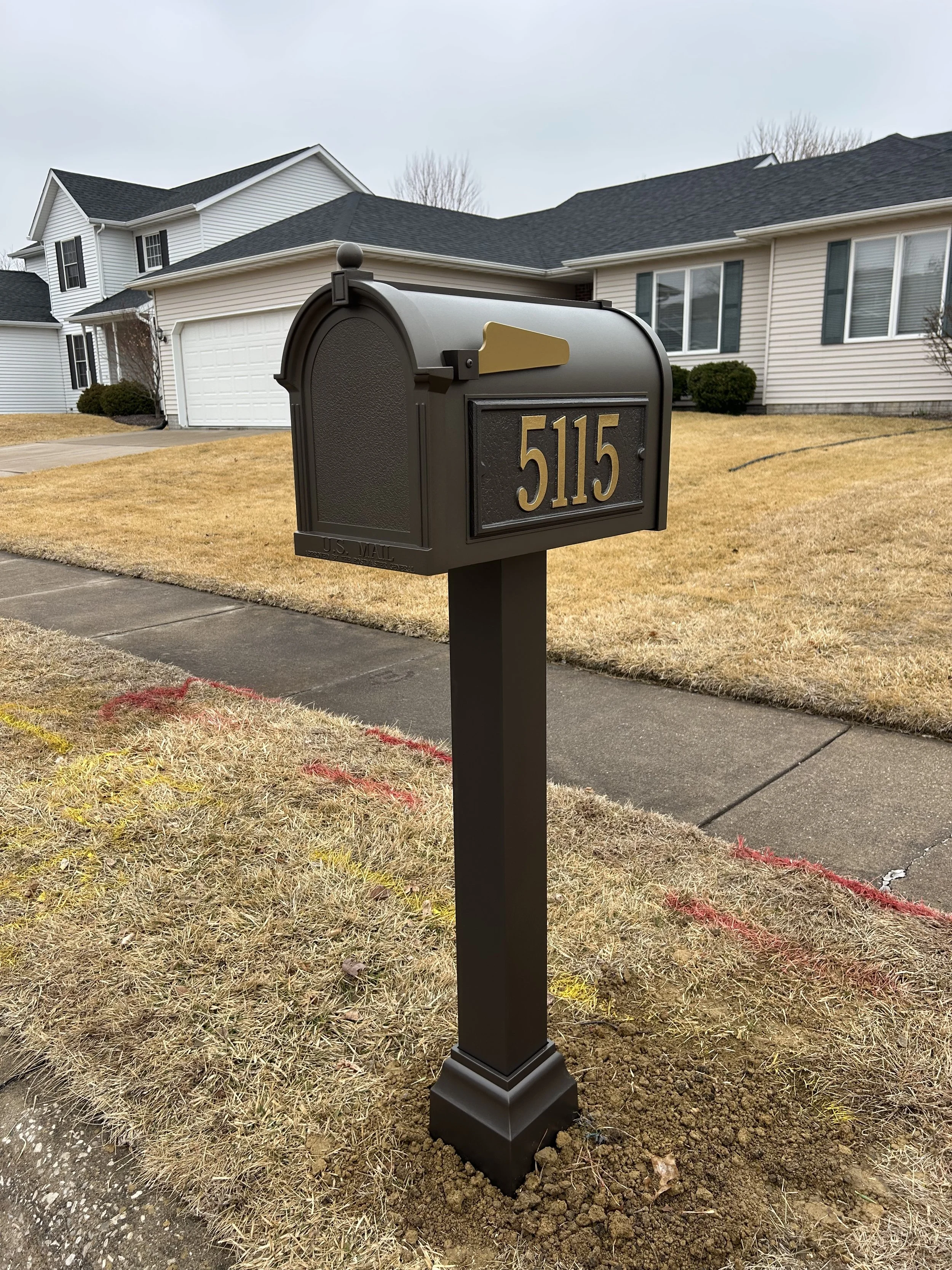 Black mailbox with gold numbers '5115' on a black post in a front yard, with a suburban house and driveway in the background.