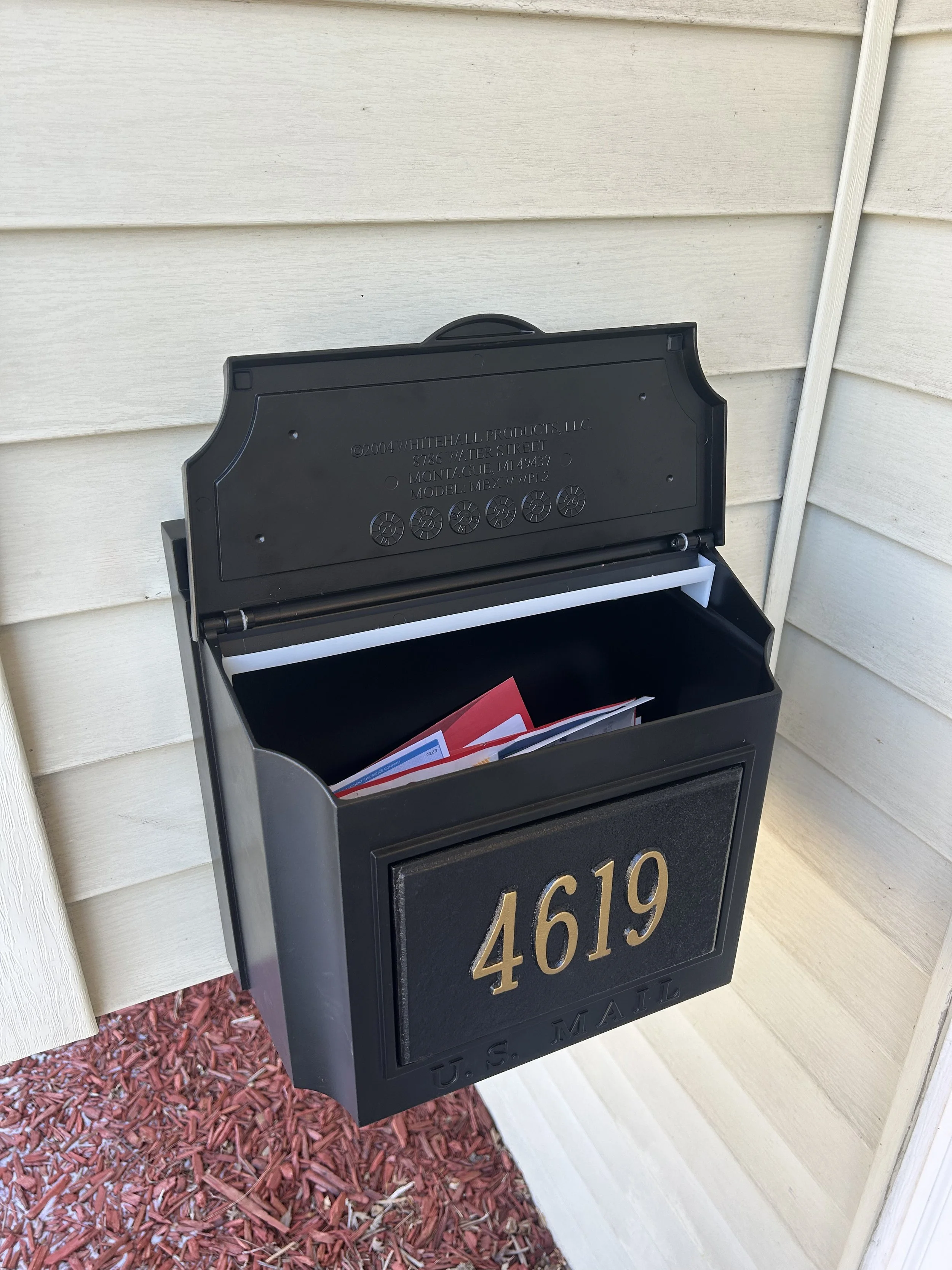 Black U.S. mailbox with the house number 4619 in gold on the front, mounted on the exterior wall of a house with beige siding, with red mulch and a small patch of grass underneath.