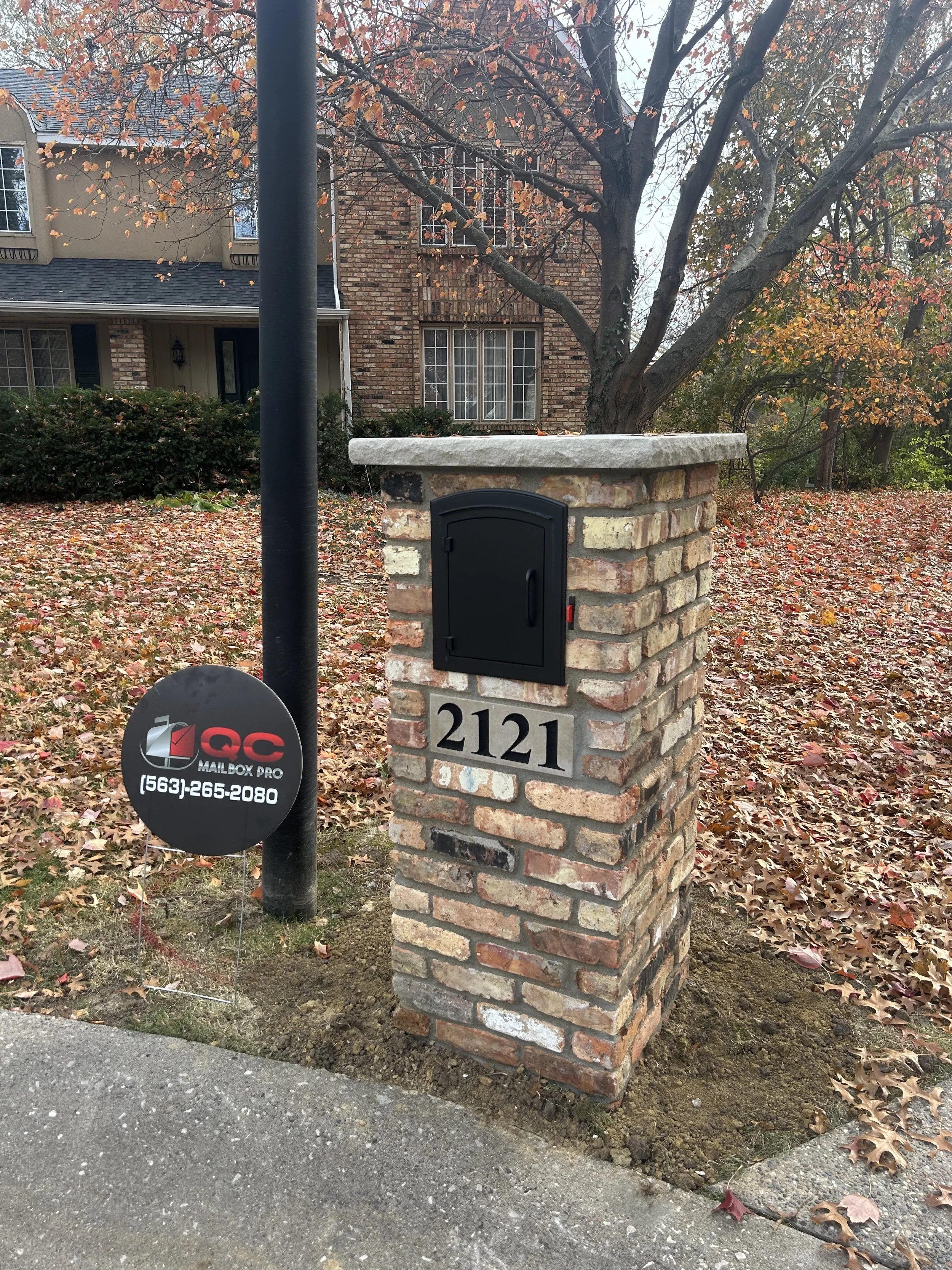 Brick mailbox with house number 2121 in front of a residential house, next to a black pole and a round black sign advertising QC Mailbox Pro with phone number 563-265-2080, during fall with leaves on the ground.