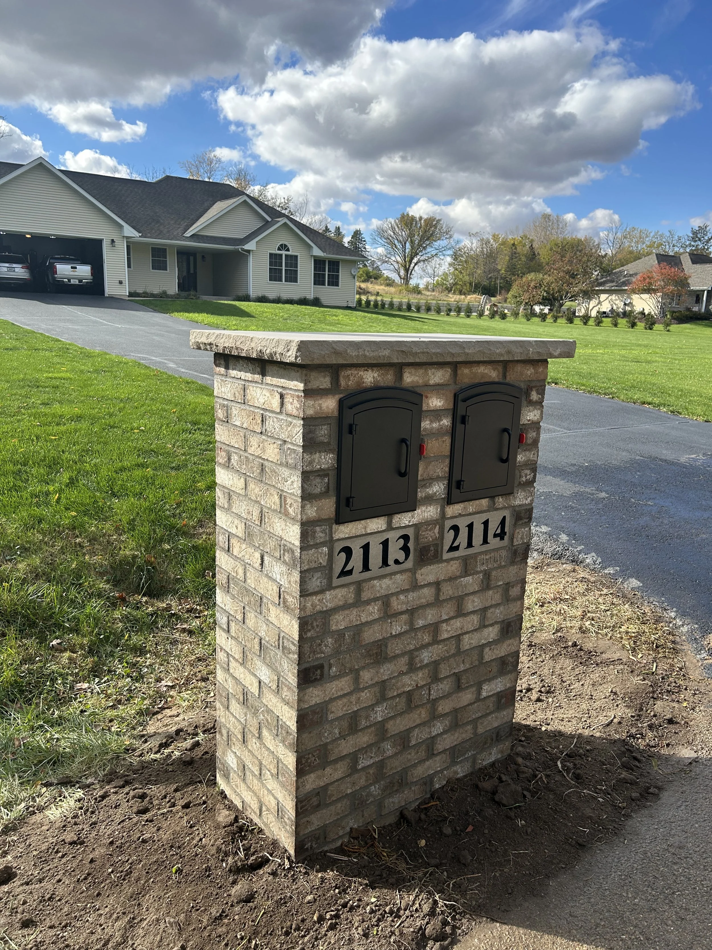 A brick mailbox post with two black mailbox doors and house numbers 2113 and 2114 on a grassy front yard under partly cloudy skies.