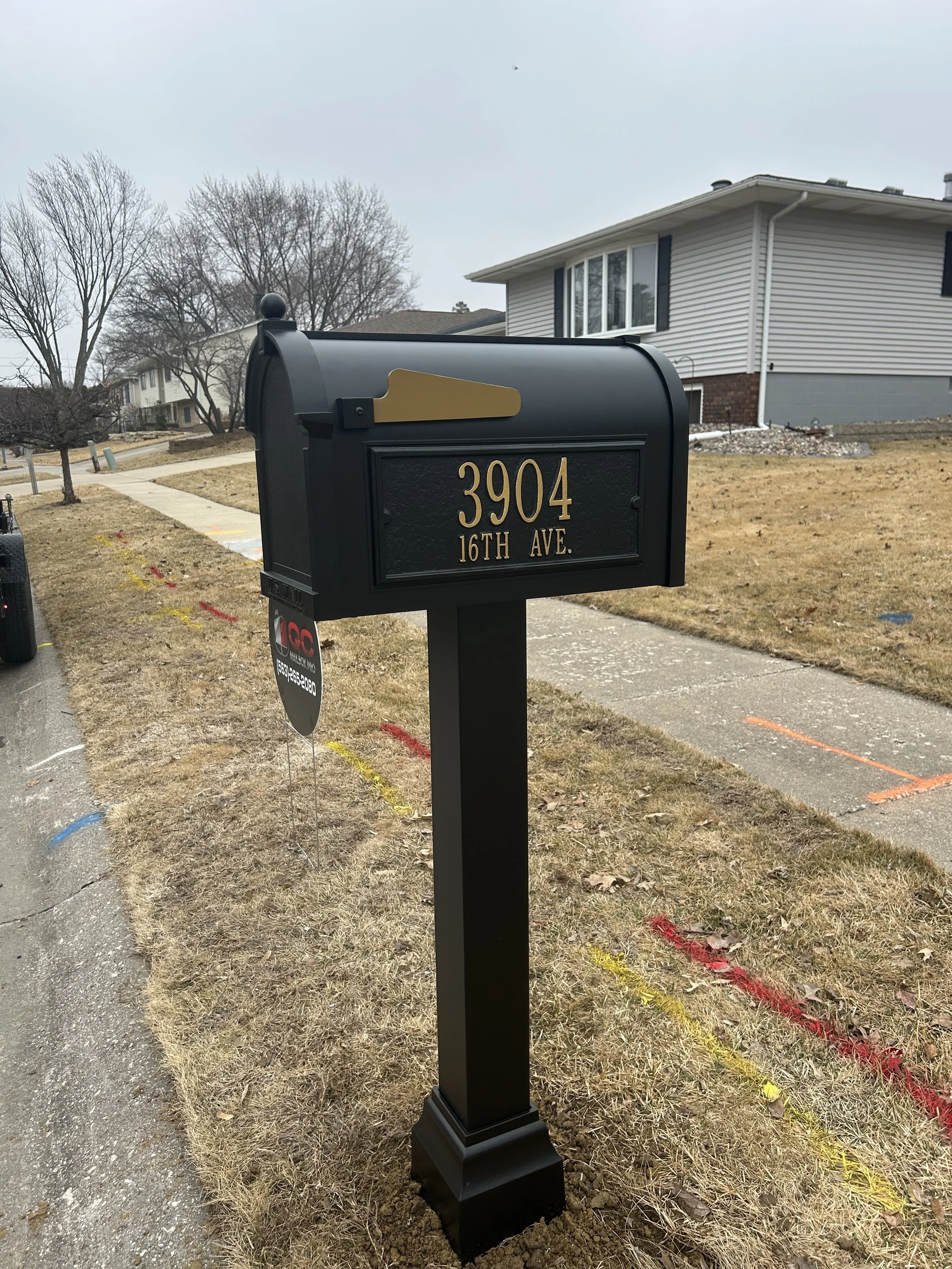 Black mailbox with address 3904 16th Ave. in gold lettering, located on a sidewalk in a residential neighborhood with a house and leafless trees in the background.