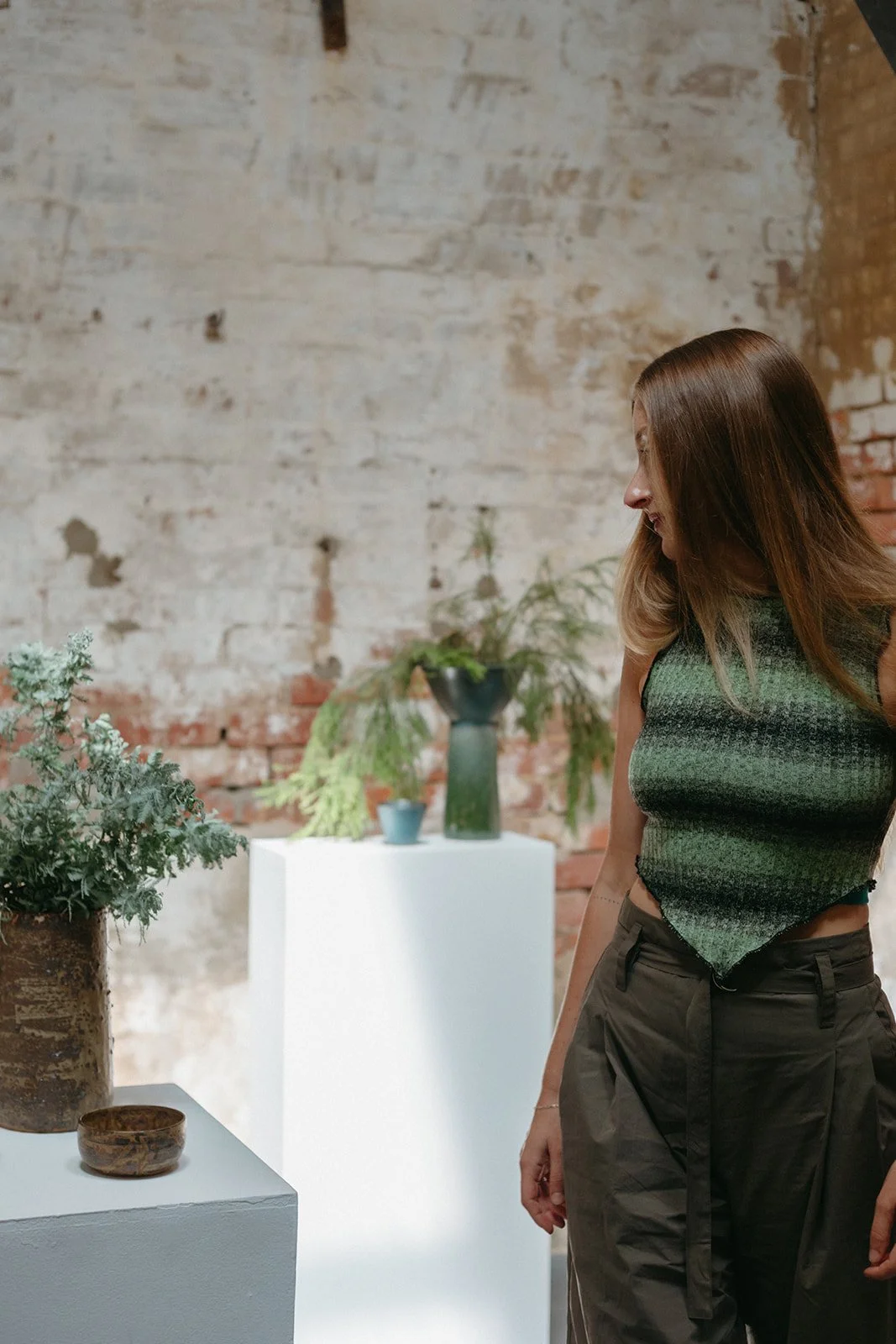 a women stands in an art gallery looking at a ceramic piece, she is wearing a green top and trousers.