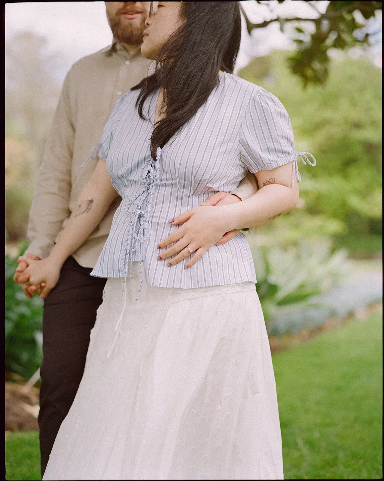 An engaged couple stand together in the park holding hands.