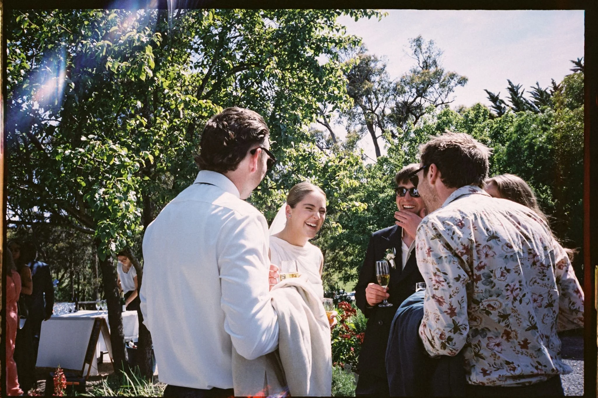 A group of young adults at a wedding, smiling and chatting with drinks in hand, surrounded by trees and greenery on a sunny day.