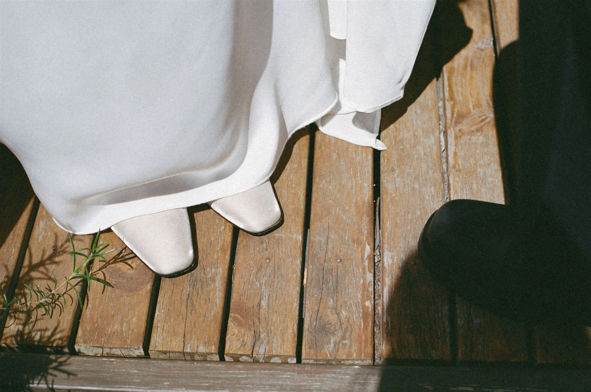 Close-up of a white satin wedding dress and white heels on a wooden floor, with a person wearing black shoes nearby.