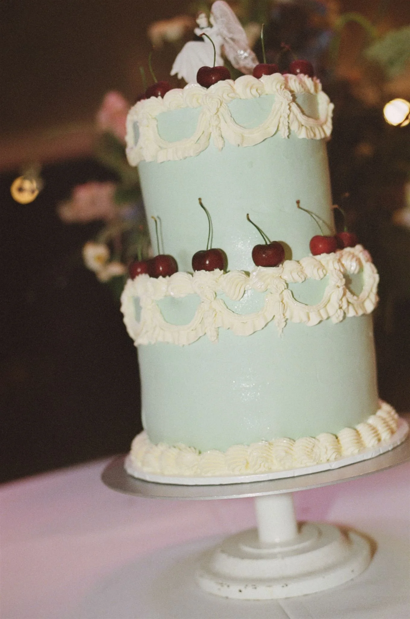 A blue tiered wedding cake with white frosting and cherries, there is a decorative couple of top. 