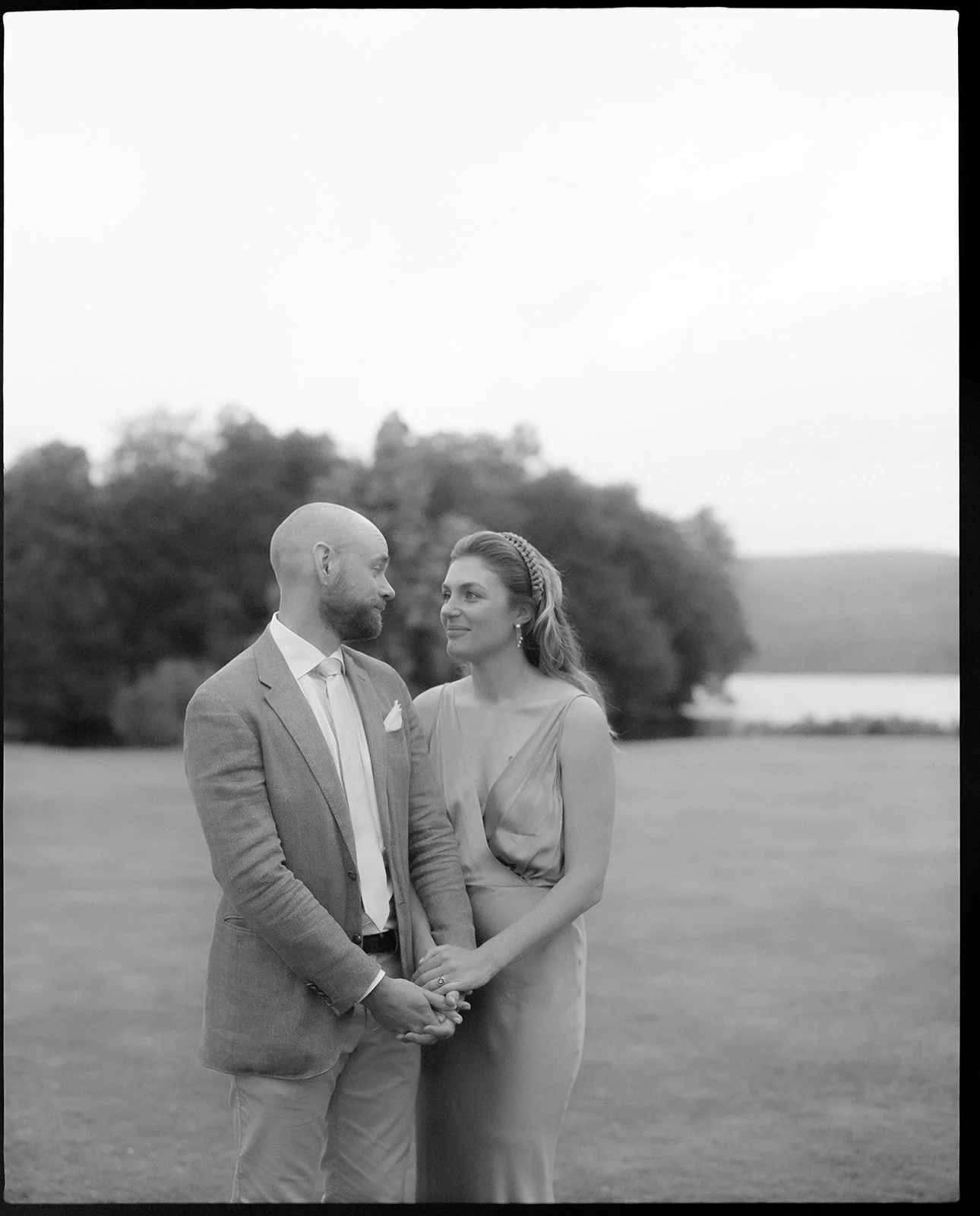 a couple stand holding hands in front of a lake, they are looking at each other and the photo is in black and white.