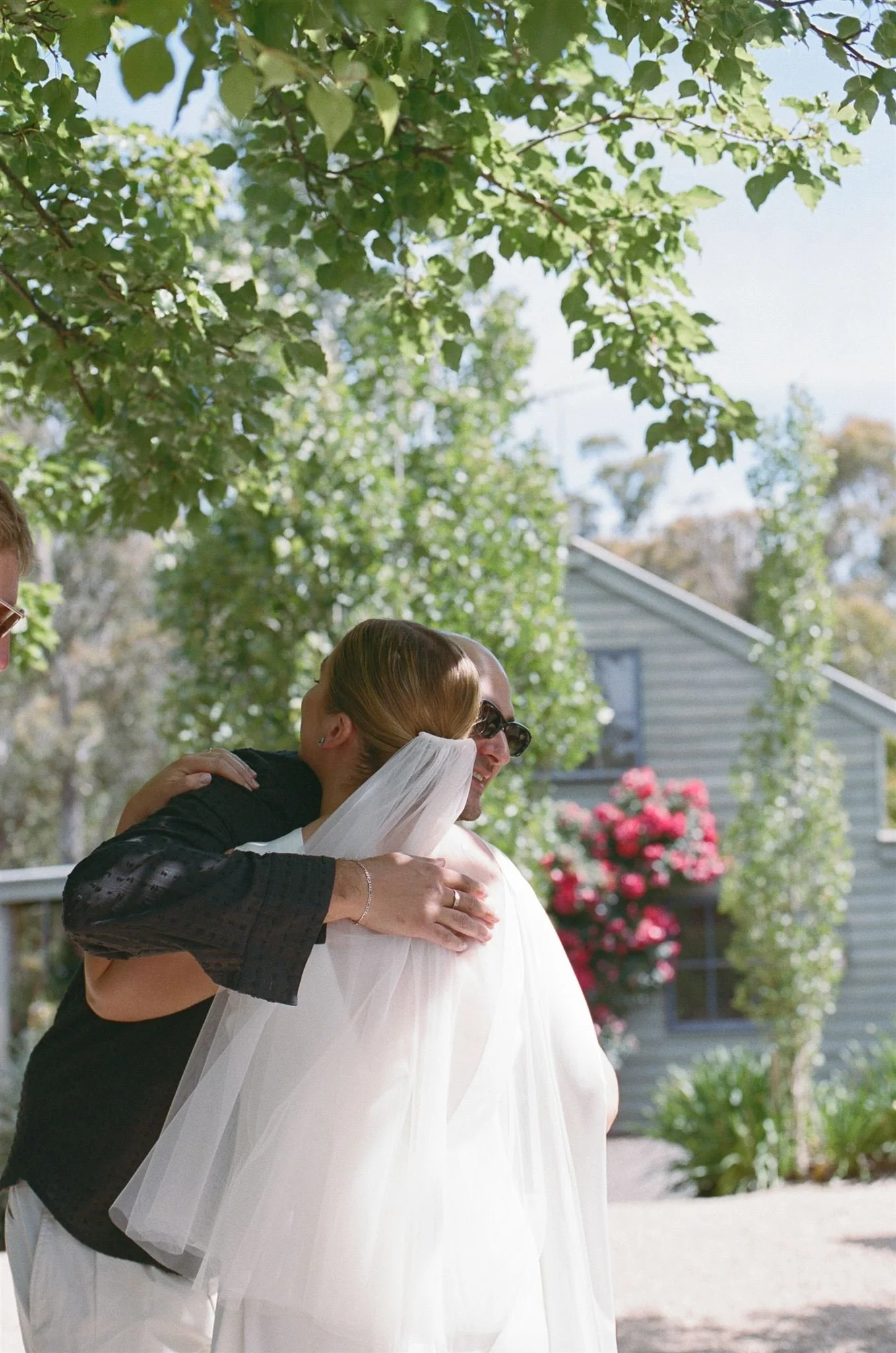 A bride hugging a guest, she has a short veil and the sun is shining through the tree's. 