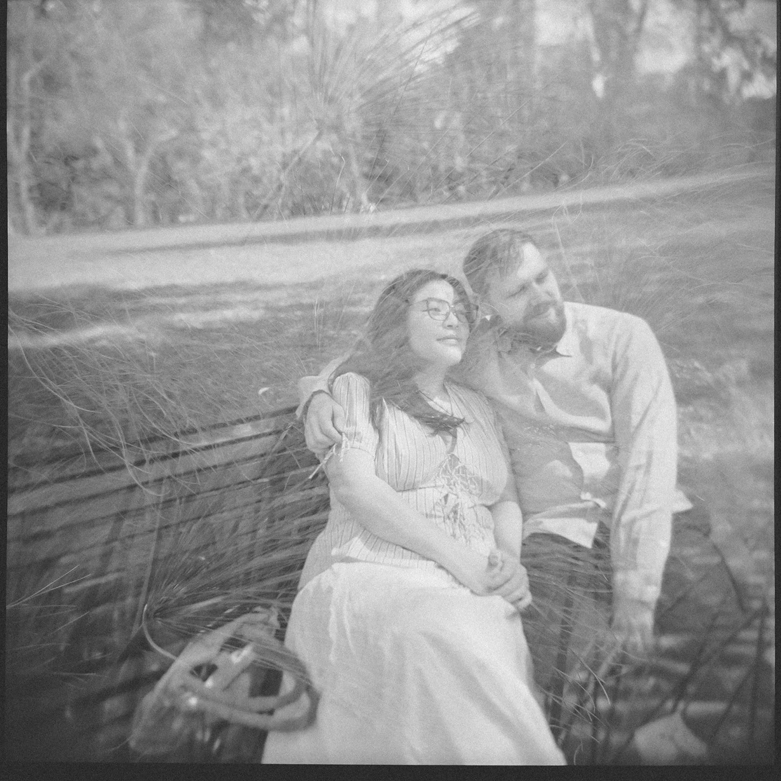 an engaged couple sit on a bench, the mans arm is around the women's shoulder, they look out to the park around them.