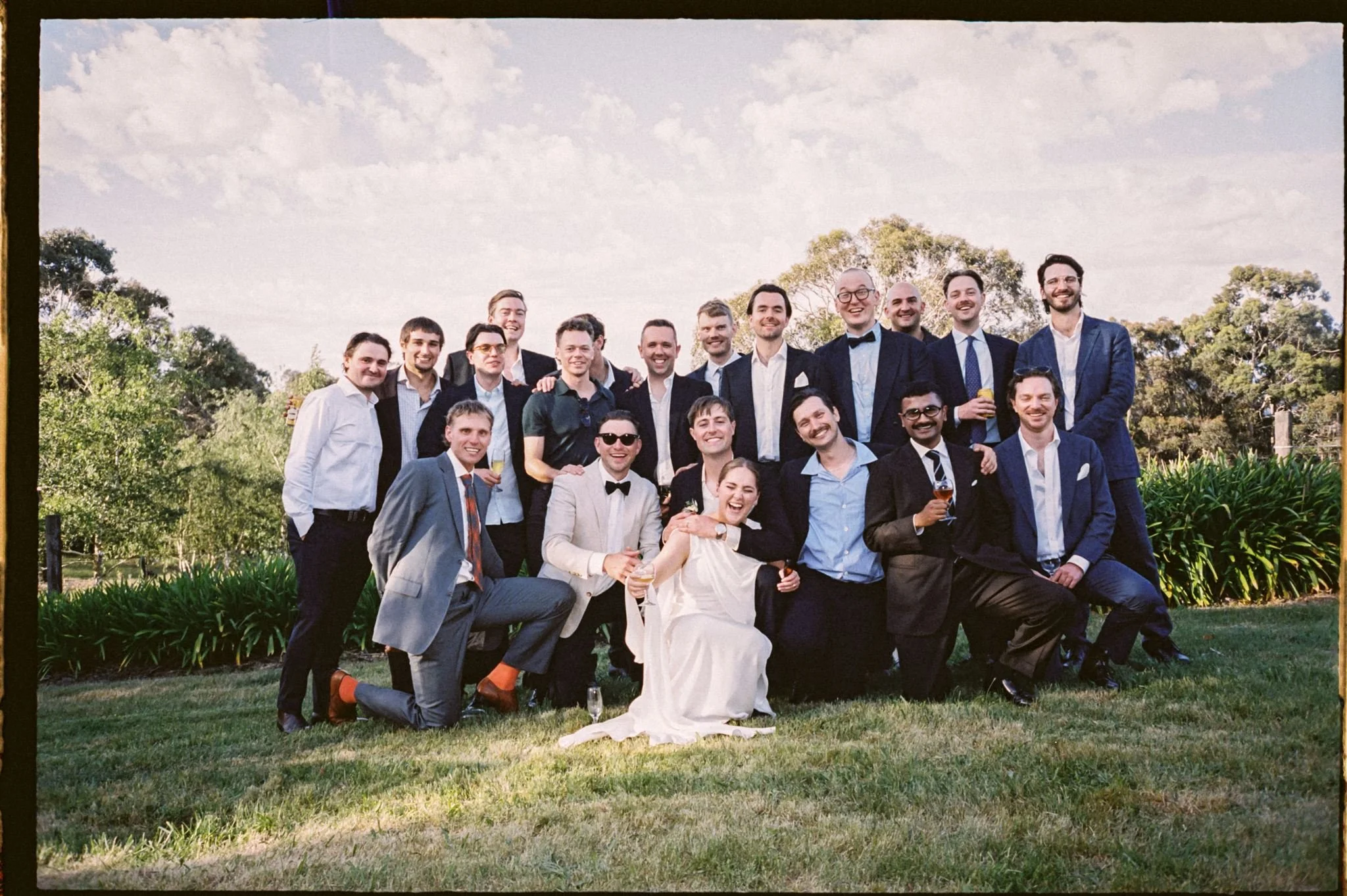 A group of people dressed in formal attire, posing outdoors on a grassy area with trees and a partly cloudy sky in the background.