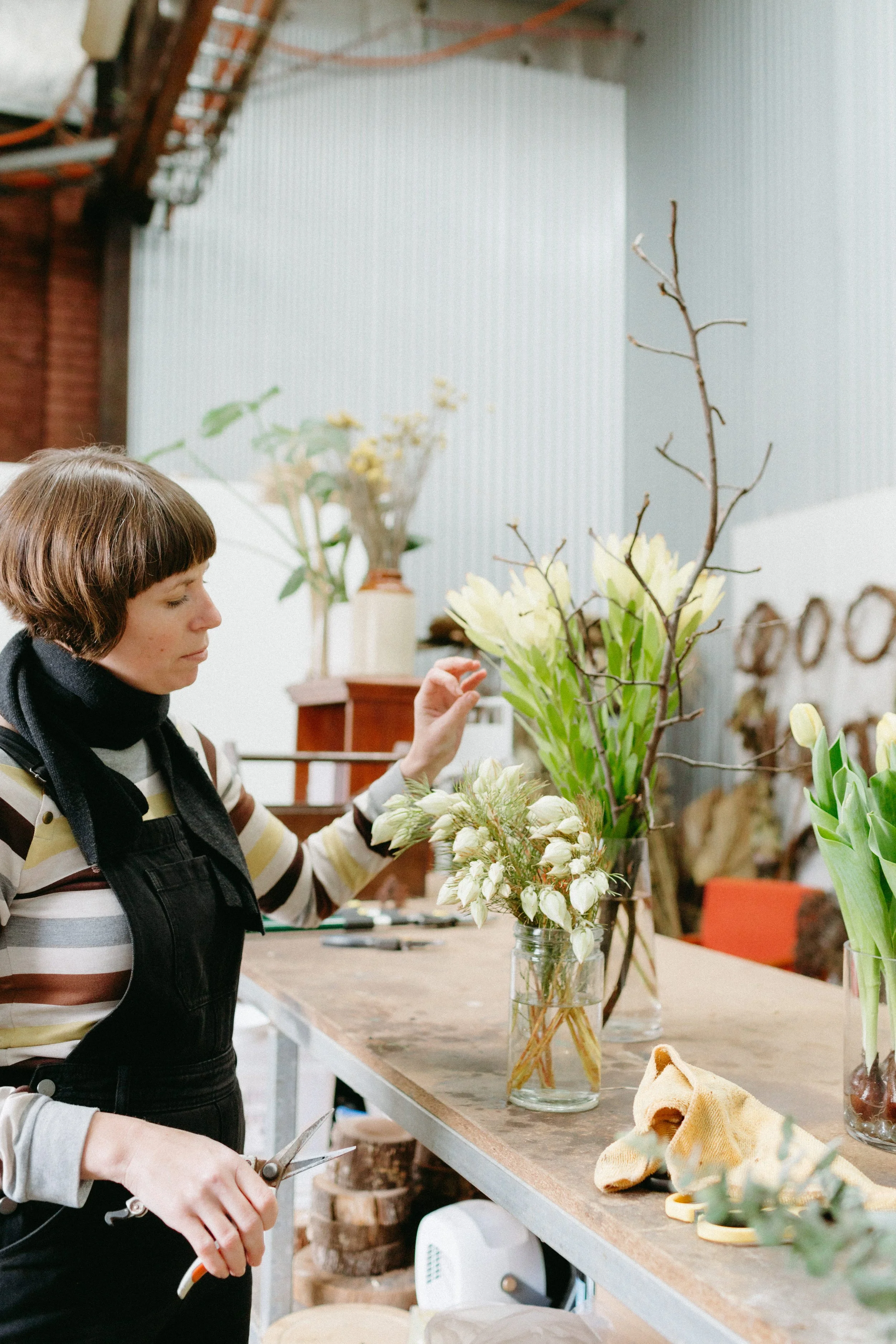 a women is arranging flowers in a workshop, she is wearing dark dungarees with a striped top and it adjusting some green and light yellow flowers.