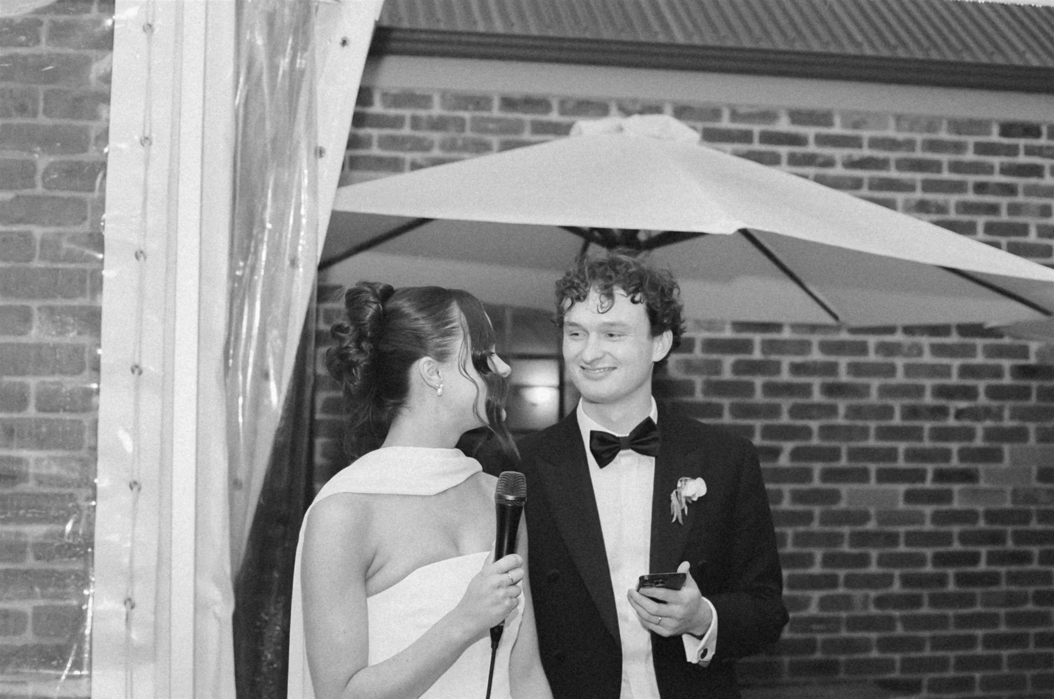 A bride and groom are giving a speech at their wedding reception. The groom is wearing a black tuxedo with bow tie and the bride is wearing a strapless dress with silk scarf. The photo is in black and white.