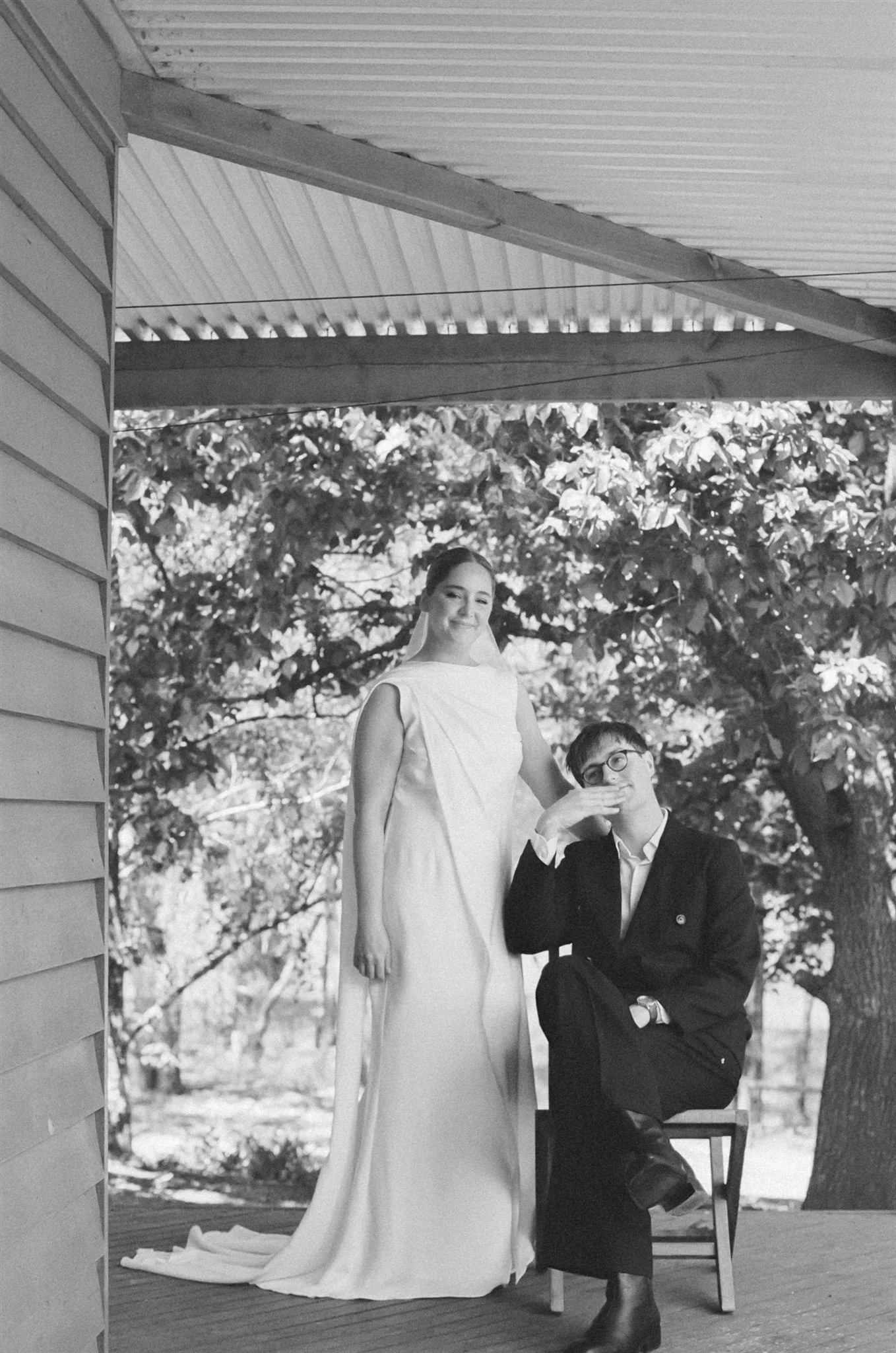 Black and white photo of a woman in a wedding dress and a man in a suit with glasses, outdoors on a porch with trees in the background.