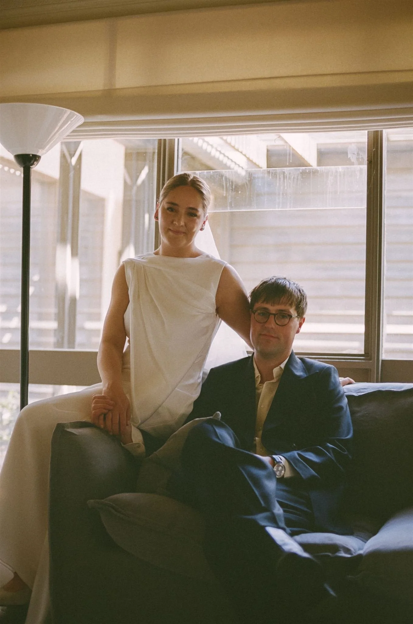 A bride in a white dress and a groom in a suit sitting on a black couch indoors near a large window with stairs outside.