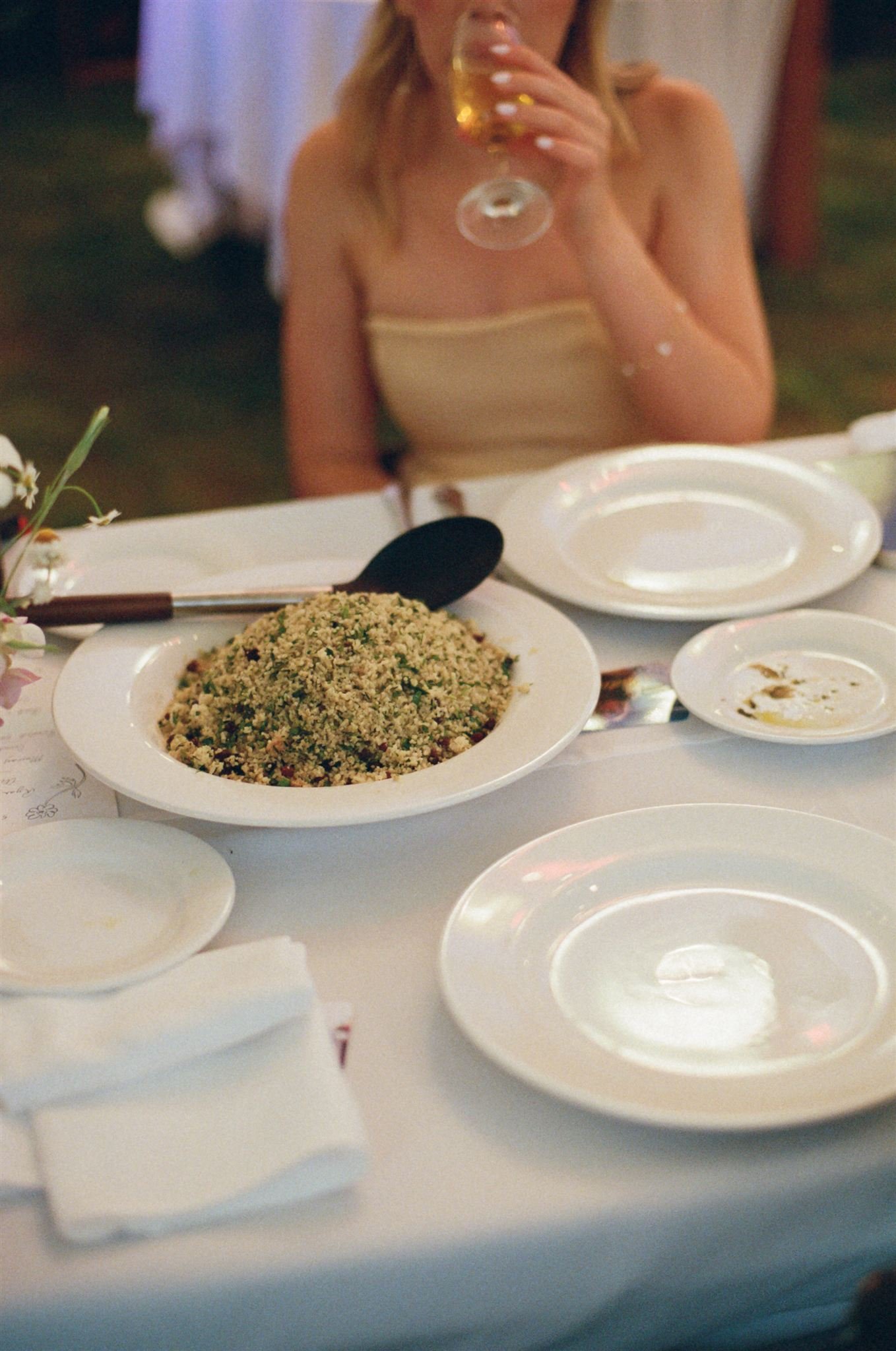 a wedding reception table with plates of food, a women sits behind it sitting a glass of wine.