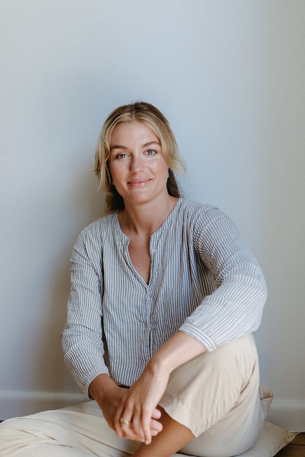 a women sits with one leg crossed against a wall, she is wearing a blue striped shirt and cream pants. She is smiling.