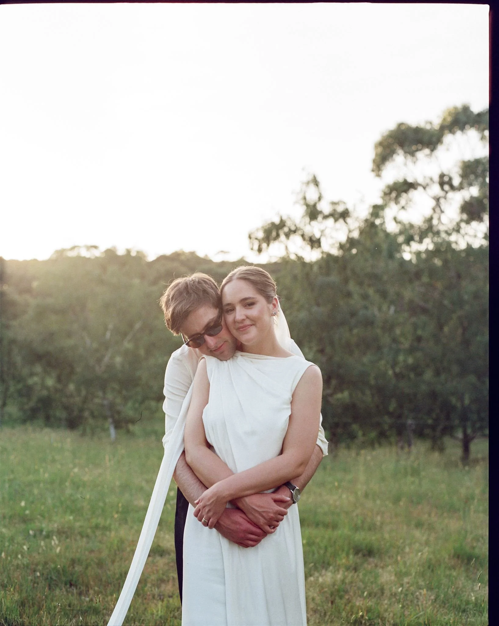 A bride and groom stand in a field with the sun setting behind them. The groom has his arms wrapped around the bride, she rests her face on his. 