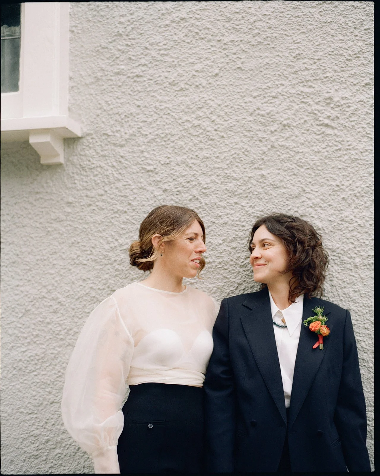 Two brides stand side by side looking at each other. They are standing in front of a wall, one bride wears a black suit jacket and the other is wearing a cream silk blouse.